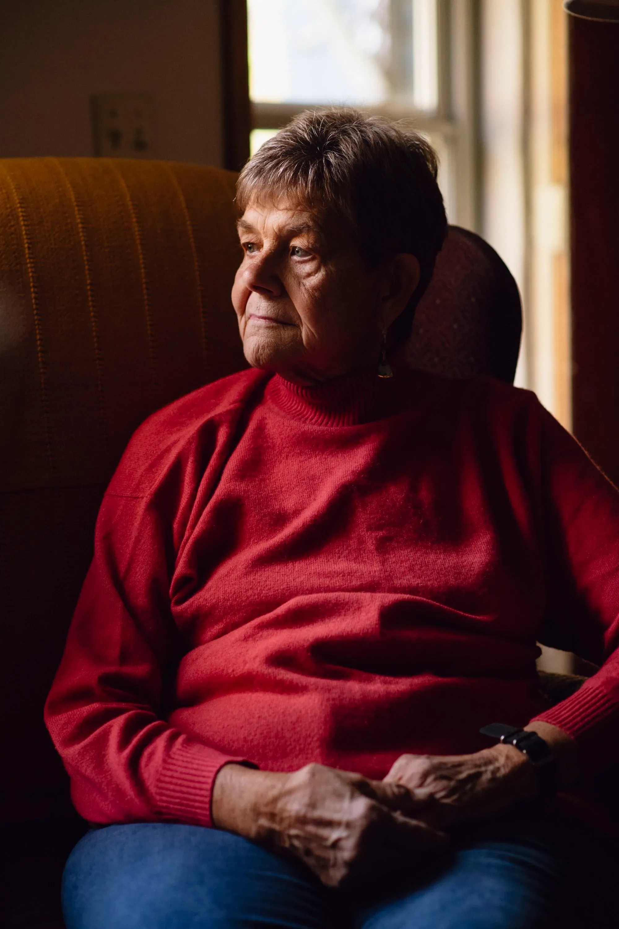 امرأة مسنّة ترتدي سترة حمراء، تجلس في غرفة مضاءة بشكل خافت، تعبر عن التأمل والهدوء، مما يعكس تأثير جائحة كوفيد-19 على المجتمع.