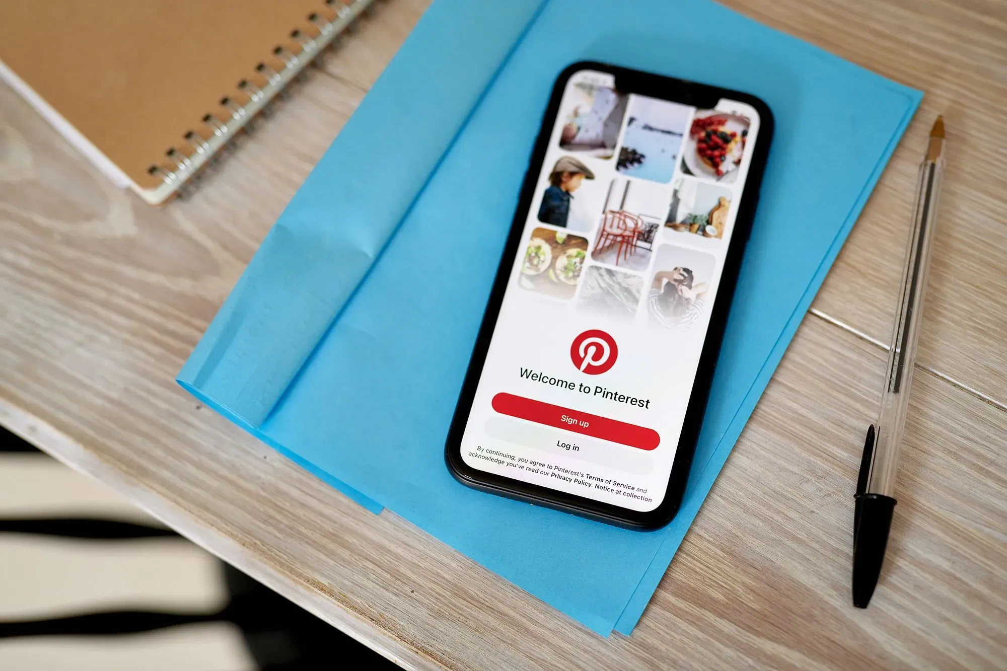 تظهر الصورة شاشة هاتف ذكي تعرض واجهة تطبيق Pinterest، مع خلفية ملونة وأدوات مكتبية مثل دفتر ملاحظات وقلم، مما يعكس تجربة المستخدم في البحث عن الإلهام.