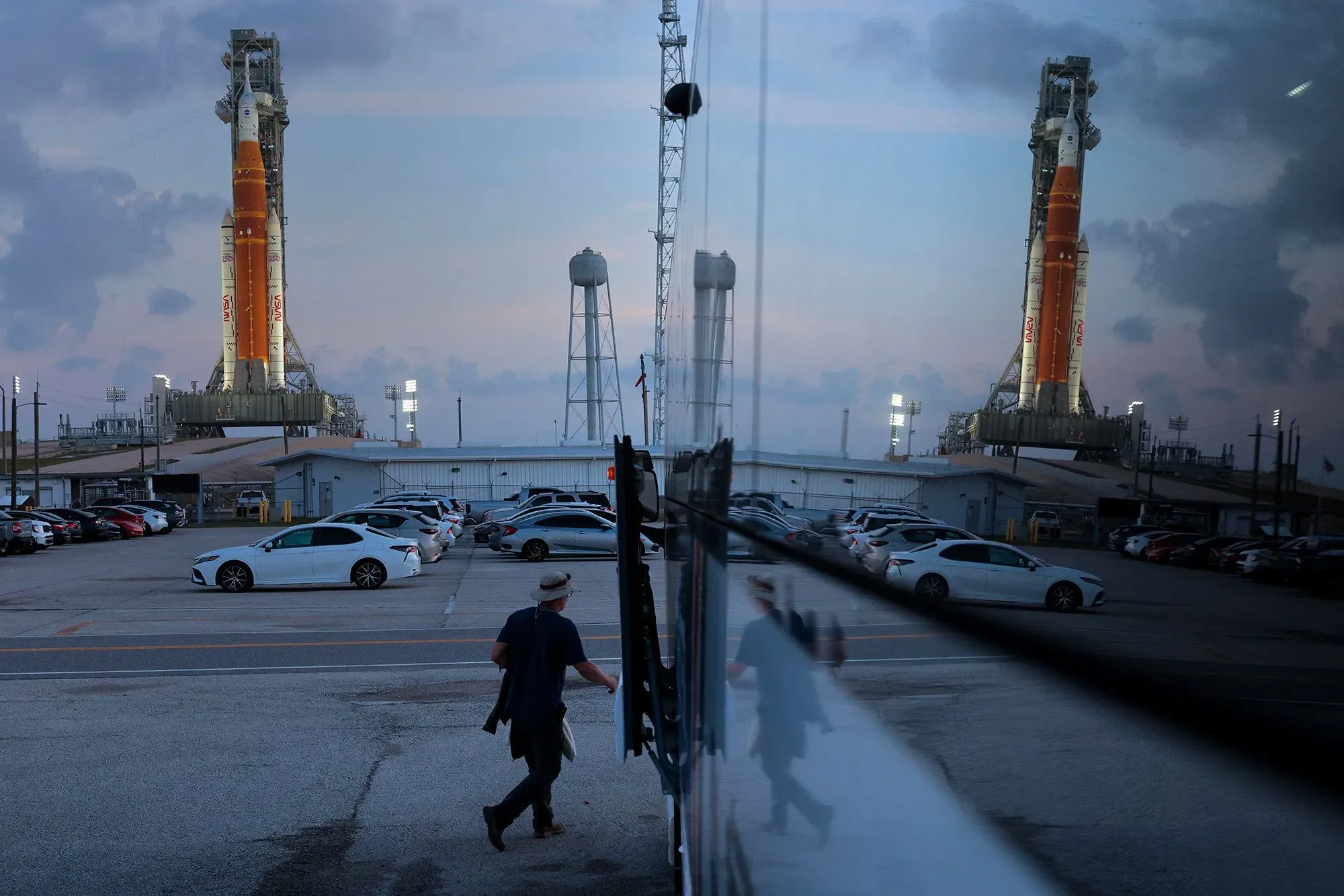 صورة تظهر صاروخ نظام الإطلاق الفضائي SLS في مركز كينيدي للفضاء، مع وجود شخص يسير بالقرب من السيارات في موقف السيارات.