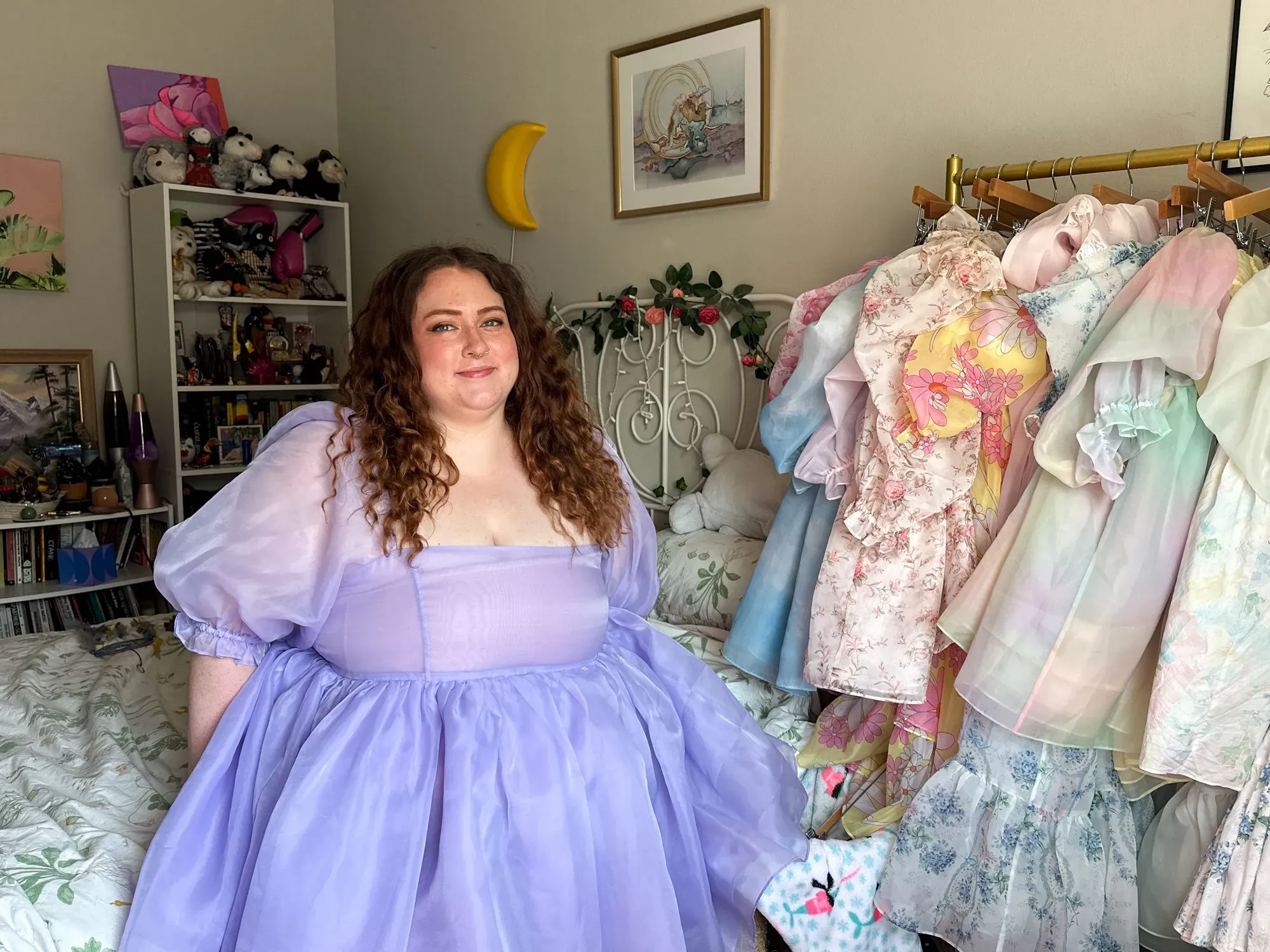 امرأة ذات مقاس كبير ترتدي فستاناً بنفسجياً في غرفة مزينة بملابس ملونة، تعكس تحديات تسوق الملابس ذات المقاسات الكبيرة.