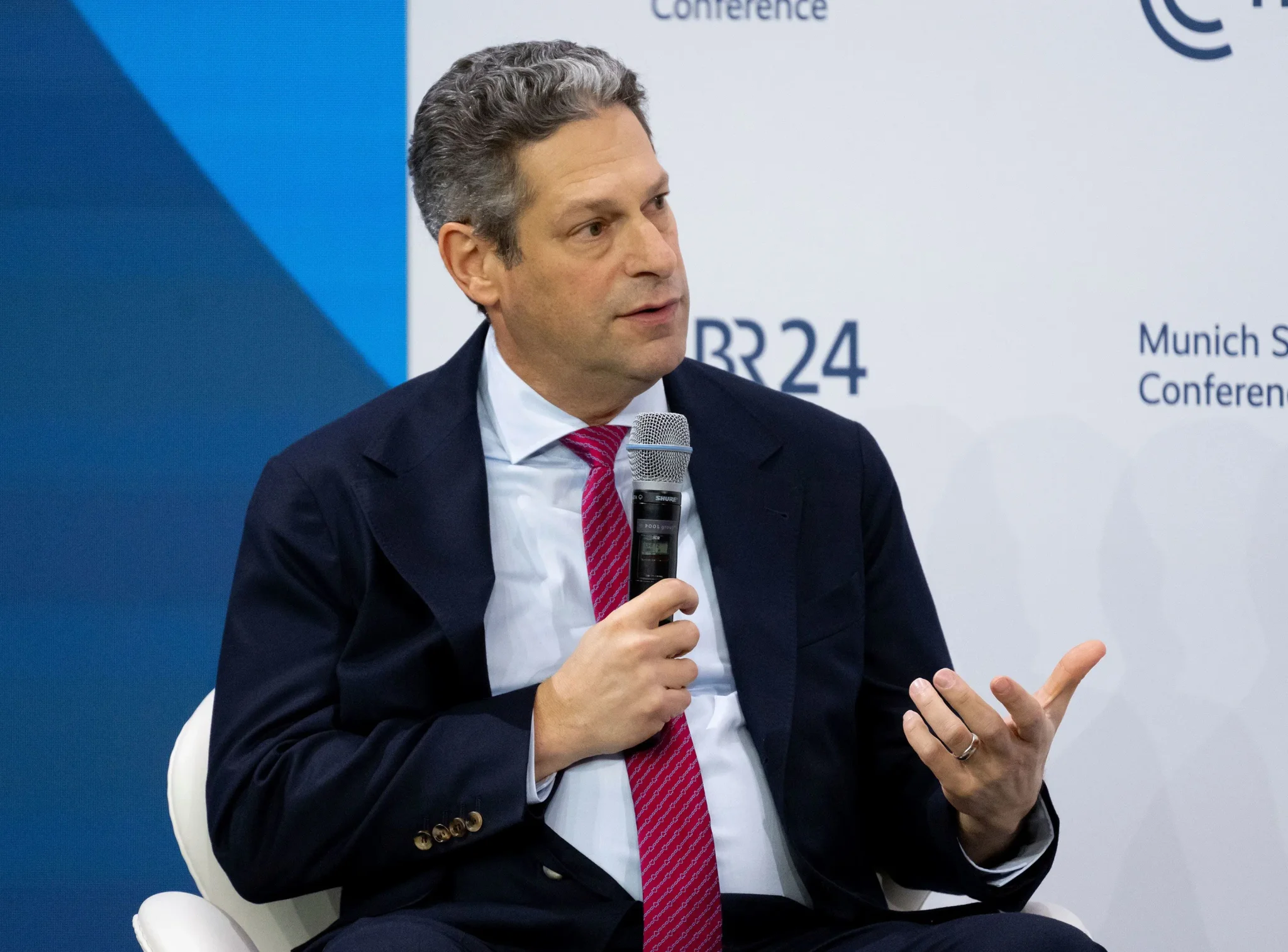 رجل يتحدث في مؤتمر ميونيخ للأمن عام 2023، مرتدياً بدلة وربطة عنق حمراء، مع التركيز على دوره في مناقشات السياسات التقنية والأمنية.