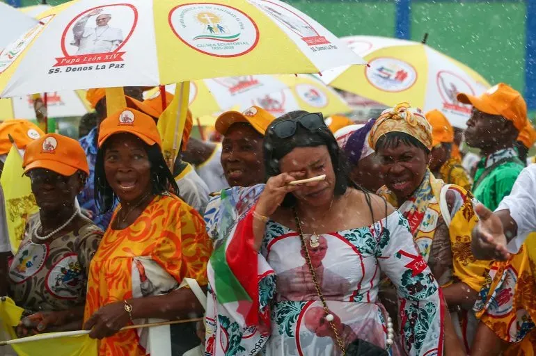 حشد من الناس يرتدون قبعات برتقالية ويحملون مظلات تحمل صور البابا، يعبرون عن مشاعرهم أثناء المطر في حدث ديني في غينيا الاستوائية.