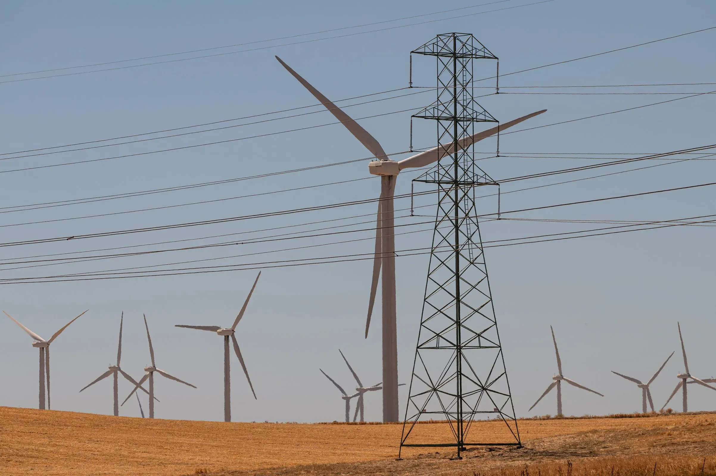 أبراج كهرباء عالية مع توربينات رياح في الخلفية، تعكس التحديات في قطاع الطاقة الأمريكية وارتفاع تكاليف الكهرباء.