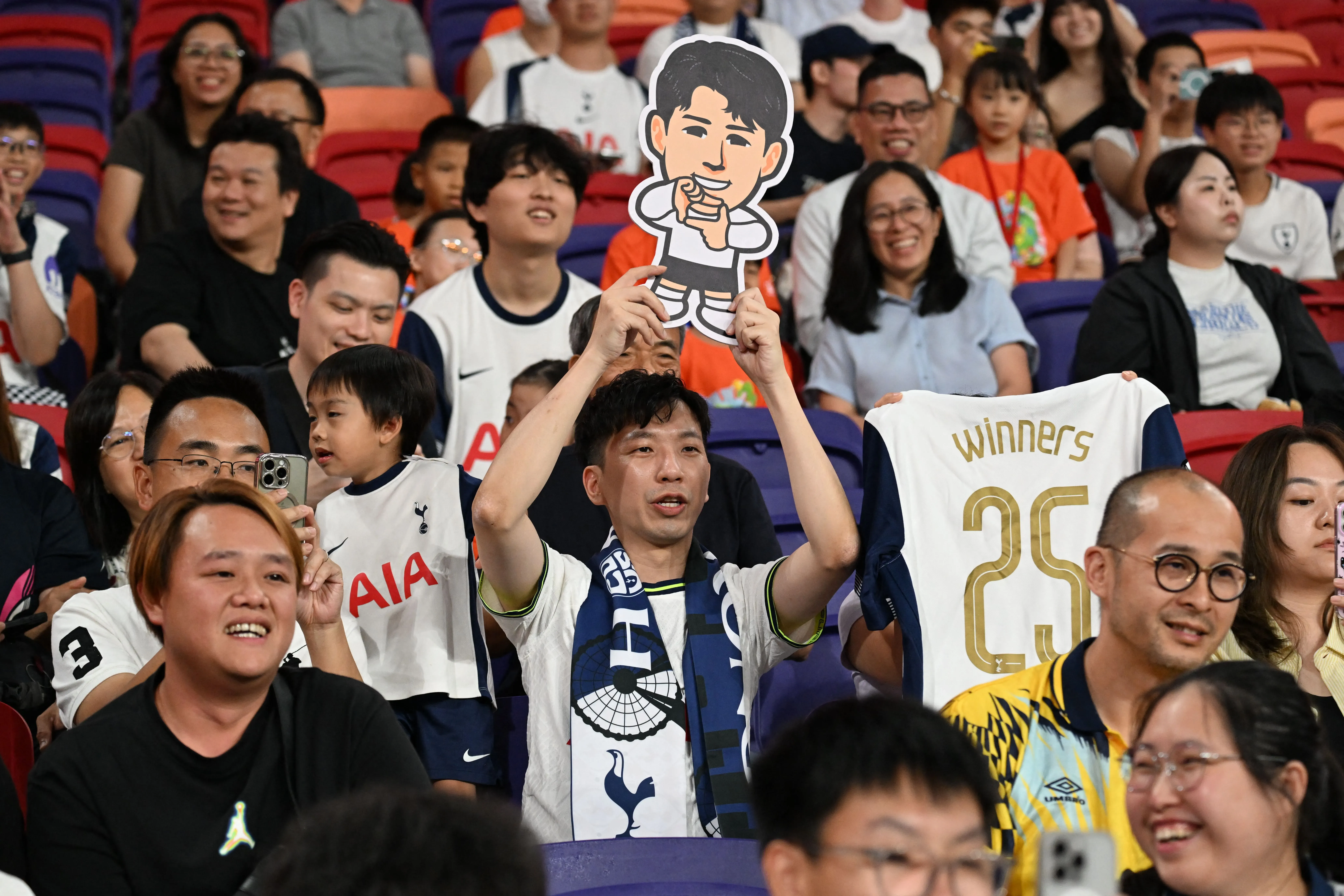 جماهير توتنهام هوتسبير في ملعب كاي تاك بهونغ كونغ، تحمل لافتة لشخصية كرتونية، تعكس حماسهم لمباراة ديربي شمال لندن.