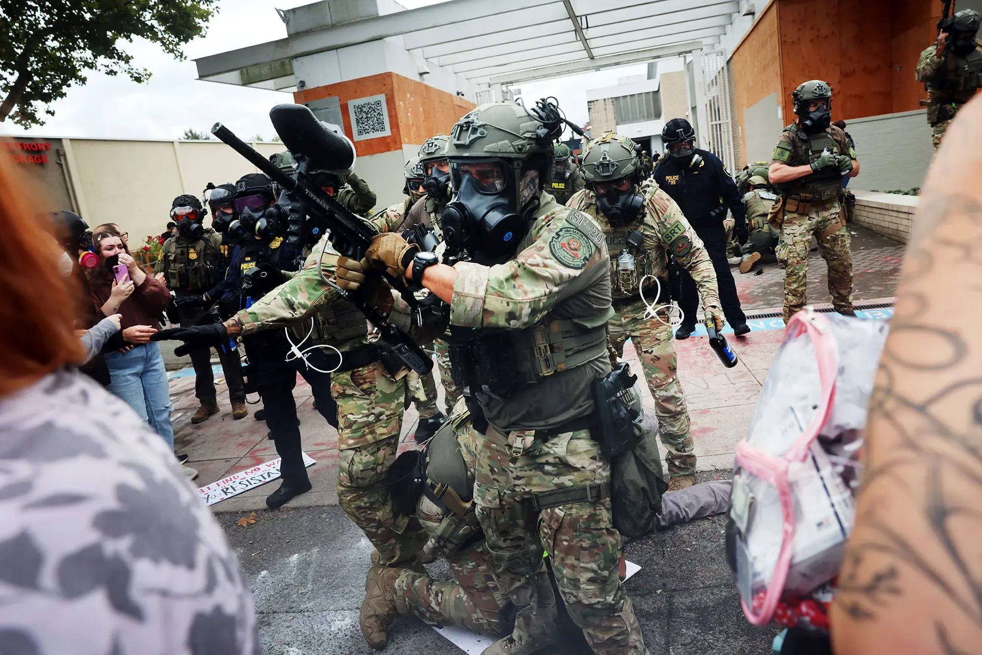 تظهر الصورة قوات الأمن ترتدي معدات مكافحة الشغب أثناء تدخلهم في مظاهرة، مع وجود متظاهرين في الخلفية.