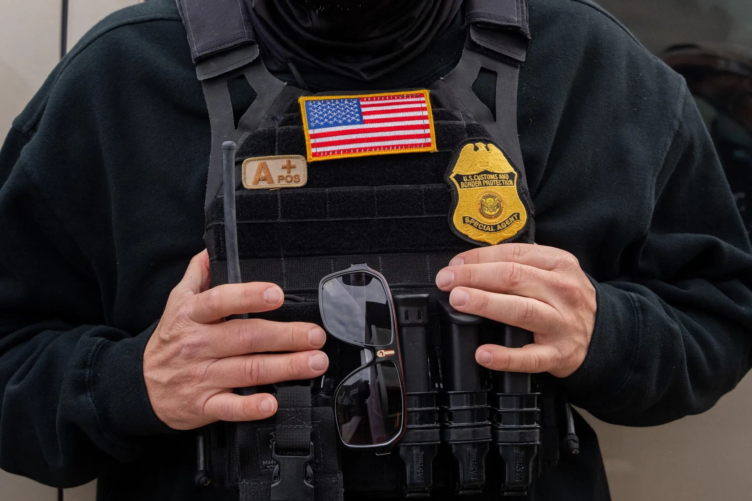 شخص يرتدي سترة واقية تحمل شعار إدارة الهجرة والجمارك الأمريكية، ممسكًا بنظارات شمسية، مما يبرز دور الوكالة في قضايا الهجرة.