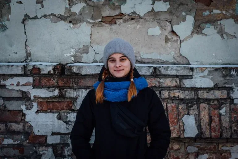 امرأة شابة ترتدي قبعة وسكارف، تقف أمام جدار متصدع في كييف، تعكس الأجواء الصعبة في ظل انقطاع الكهرباء بسبب الحرب.