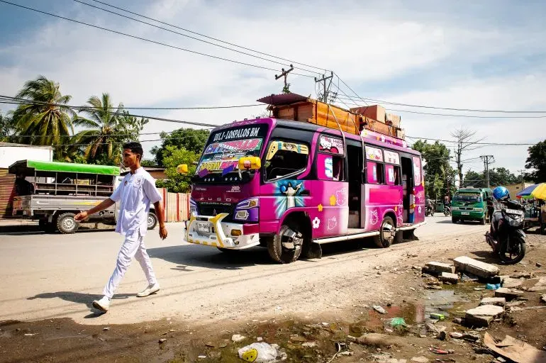 حافلة ملونة تسير في شارع مزدحم في تيمور الشرقية، مع شخص يمشي بجانبها، تعكس الحياة اليومية في البلاد بعد الاستقلال.