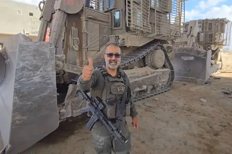 رجل يرتدي زيًا عسكريًا ويبتسم أمام جرافة عسكرية، مع سلاح في حزامه، مما يعكس أجواء الصراع في غزة.