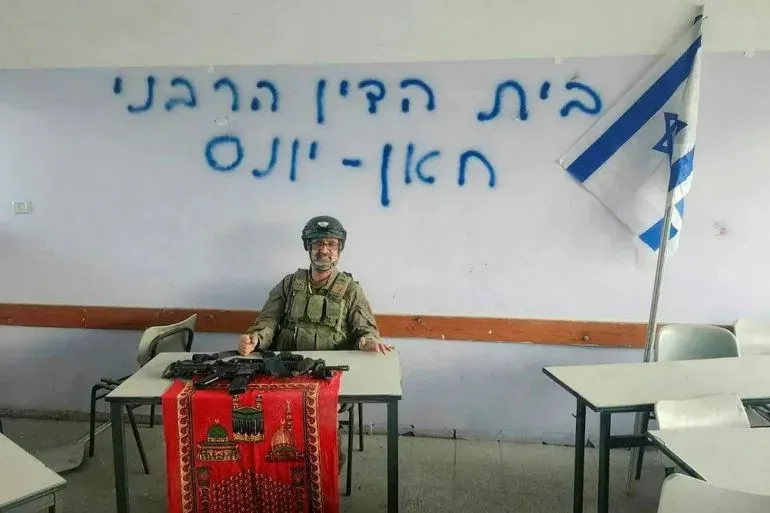 صورة لجندي إسرائيلي يجلس أمام طاولة تحمل أسلحة، في غرفة صفية تحمل علم إسرائيل وكتابات بالعبرية، تعكس التوترات في المنطقة.