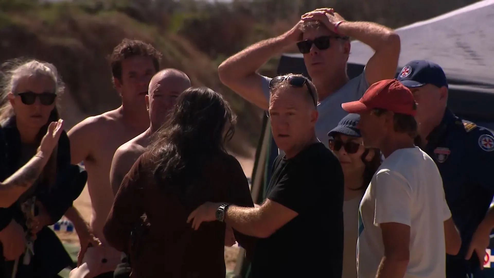 مجموعة من الأشخاص في حالة صدمة على شاطئ لونغ ريف بعد هجوم سمكة قرش أدى إلى وفاة راكب أمواج، مع وجود خدمات الطوارئ في الخلفية.