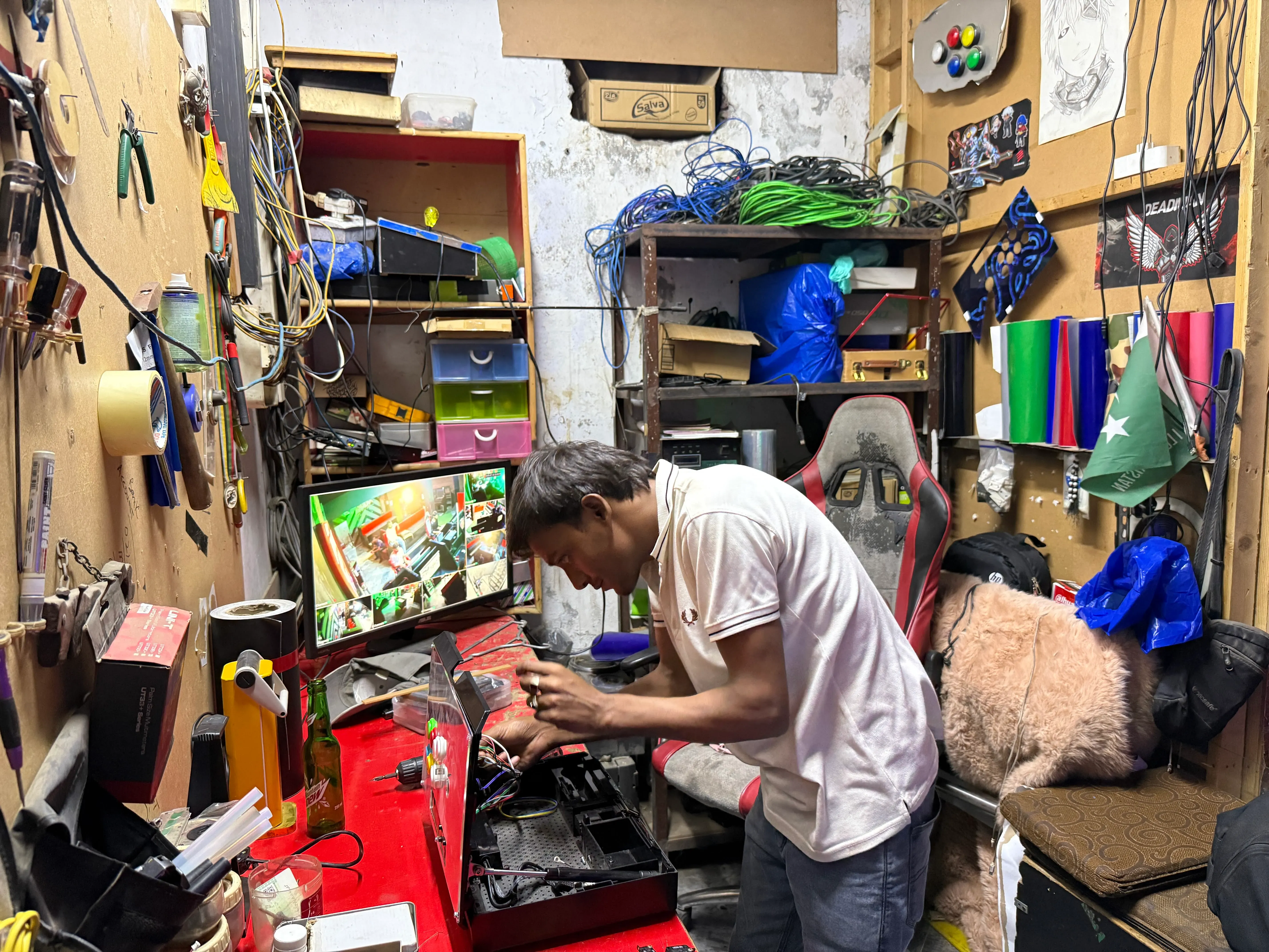 شاب يعمل على إصلاح جهاز تحكم ألعاب في ورشة صغيرة بل Lahore، محاط بأدوات ومعدات، بينما يظهر على الشاشة مشهد من لعبة Tekken.