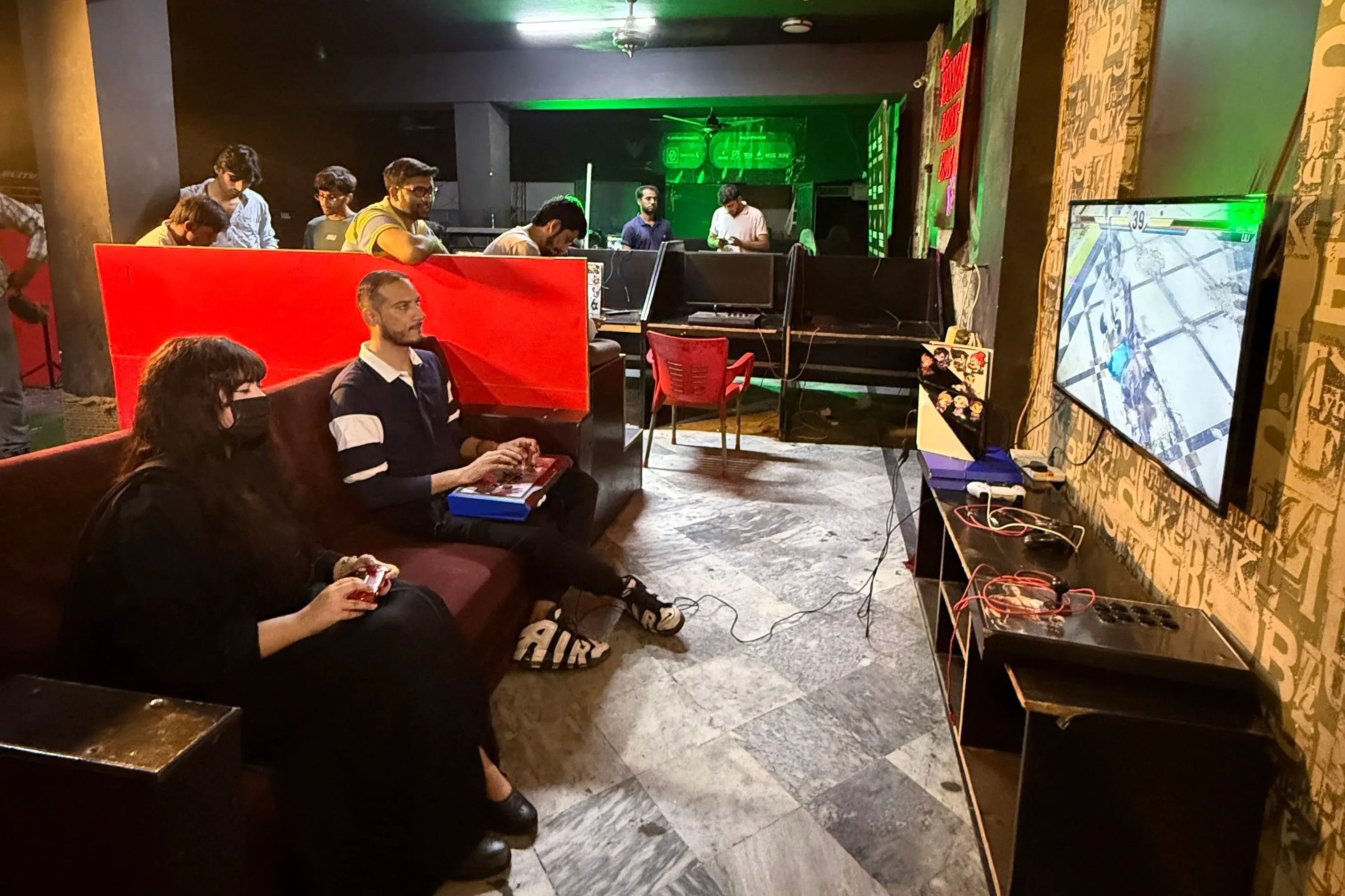 لاعبون يتنافسون في صالة ألعاب مانياكس بلهاور، حيث تُعرض لعبة Tekken على الشاشة، مع أجواء حماسية ومعدات ألعاب متوفرة.