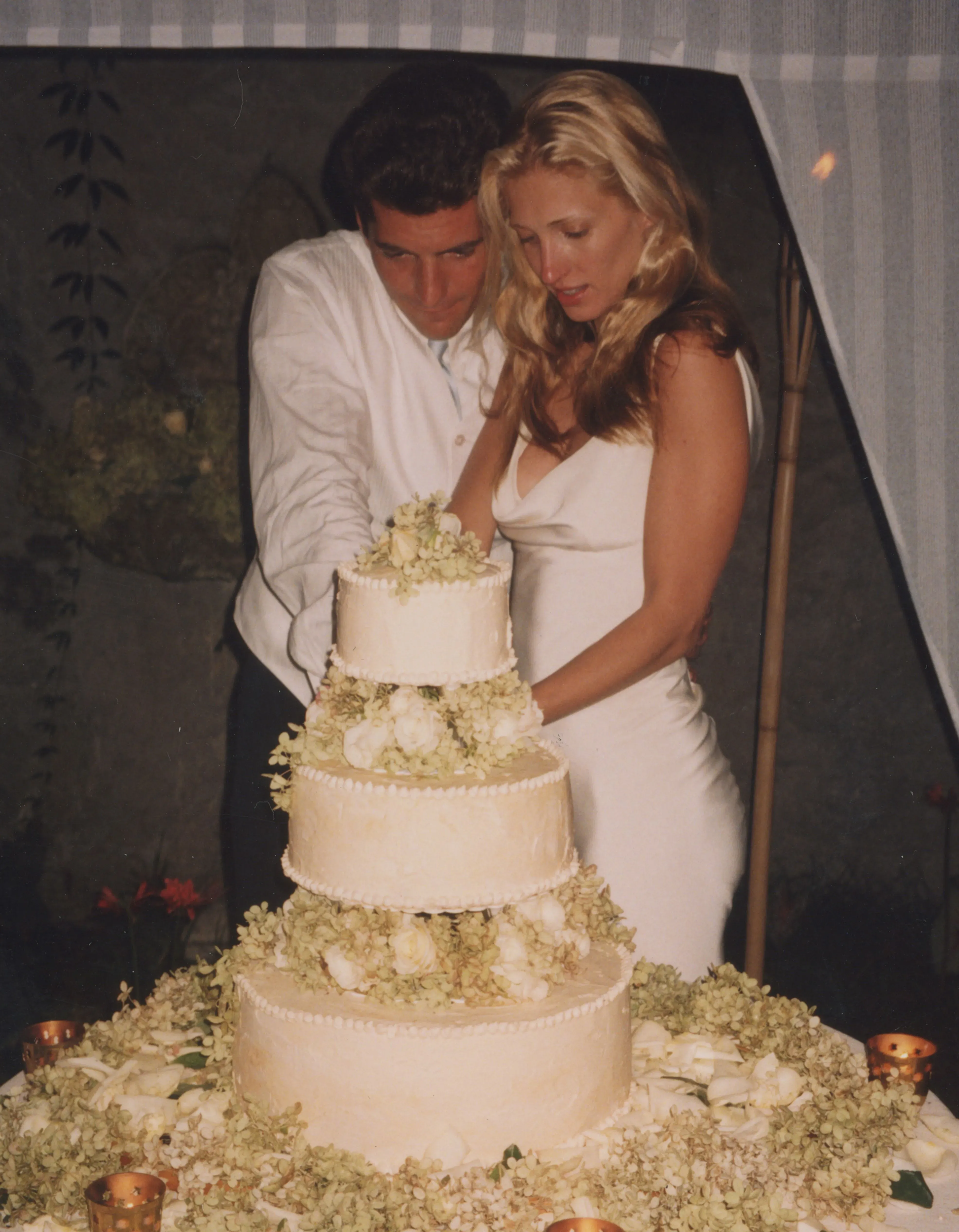 كارولين بيسيت-كينيدي وجون كينيدي الابن يقطعان كعكة زفافهما في حفل بسيط، حيث ترتدي بيسيت-كينيدي فستان زفاف أبيض أنيق من تصميم نارسيسو رودريغيز.