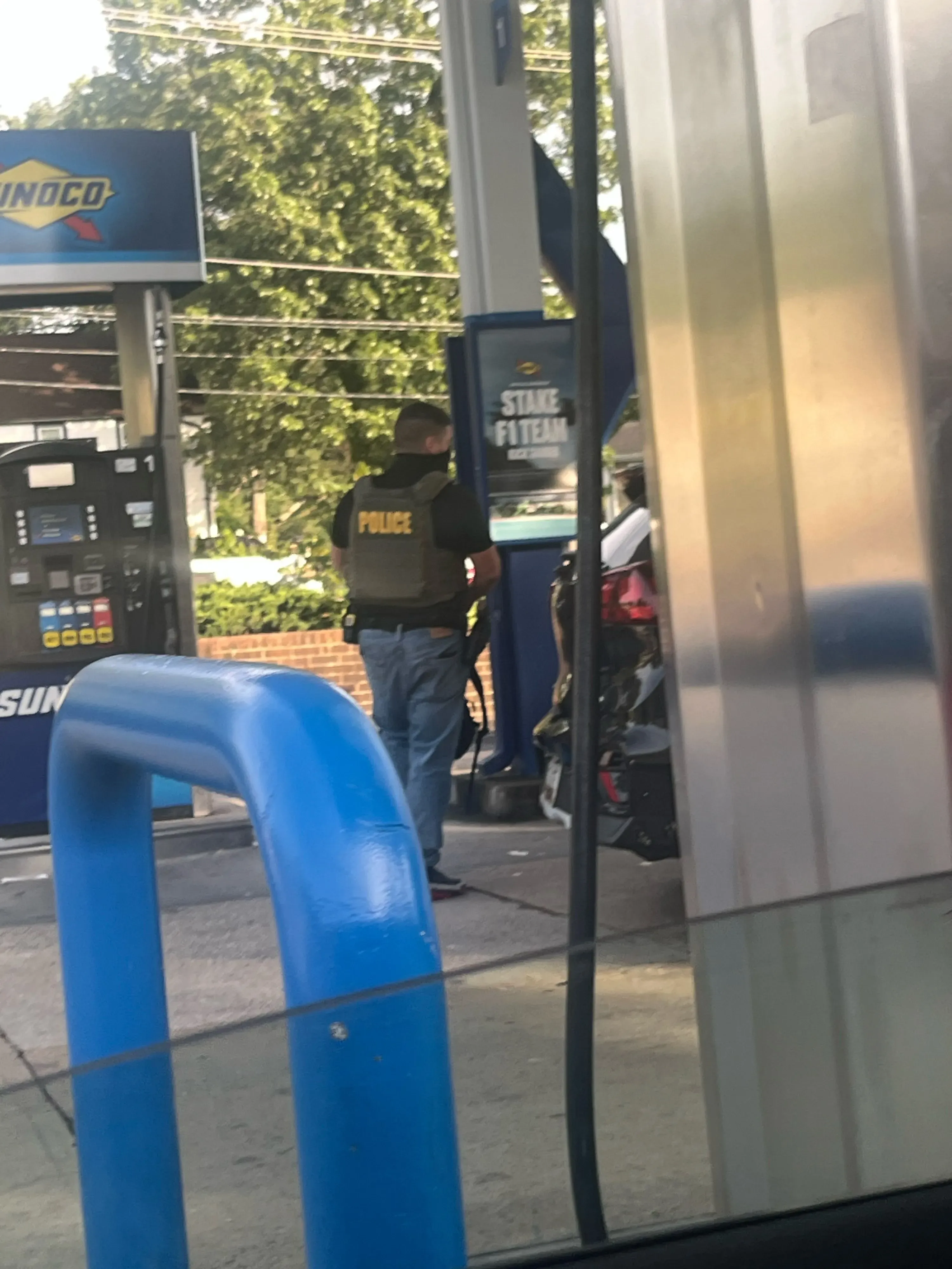 رجل يرتدي سترة تكتيكية يحمل سلاحًا في محطة وقود، بينما يتحدث مع شخص آخر يبدو مذعورًا، في مشهد يعكس توتر الاعتقال.