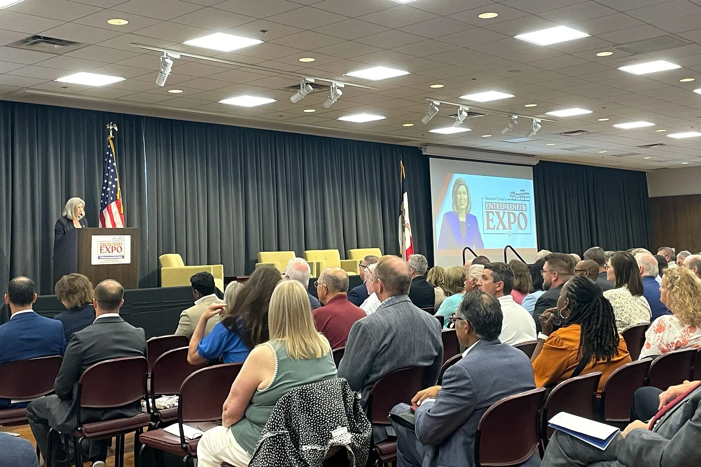 حدث معرض رواد الأعمال في ولاية أيوا، حيث تتحدث مديرة الأعمال الصغيرة كيلي لوفلر أمام جمهور من رجال الأعمال، مع وجود العلم الأمريكي في الخلفية.