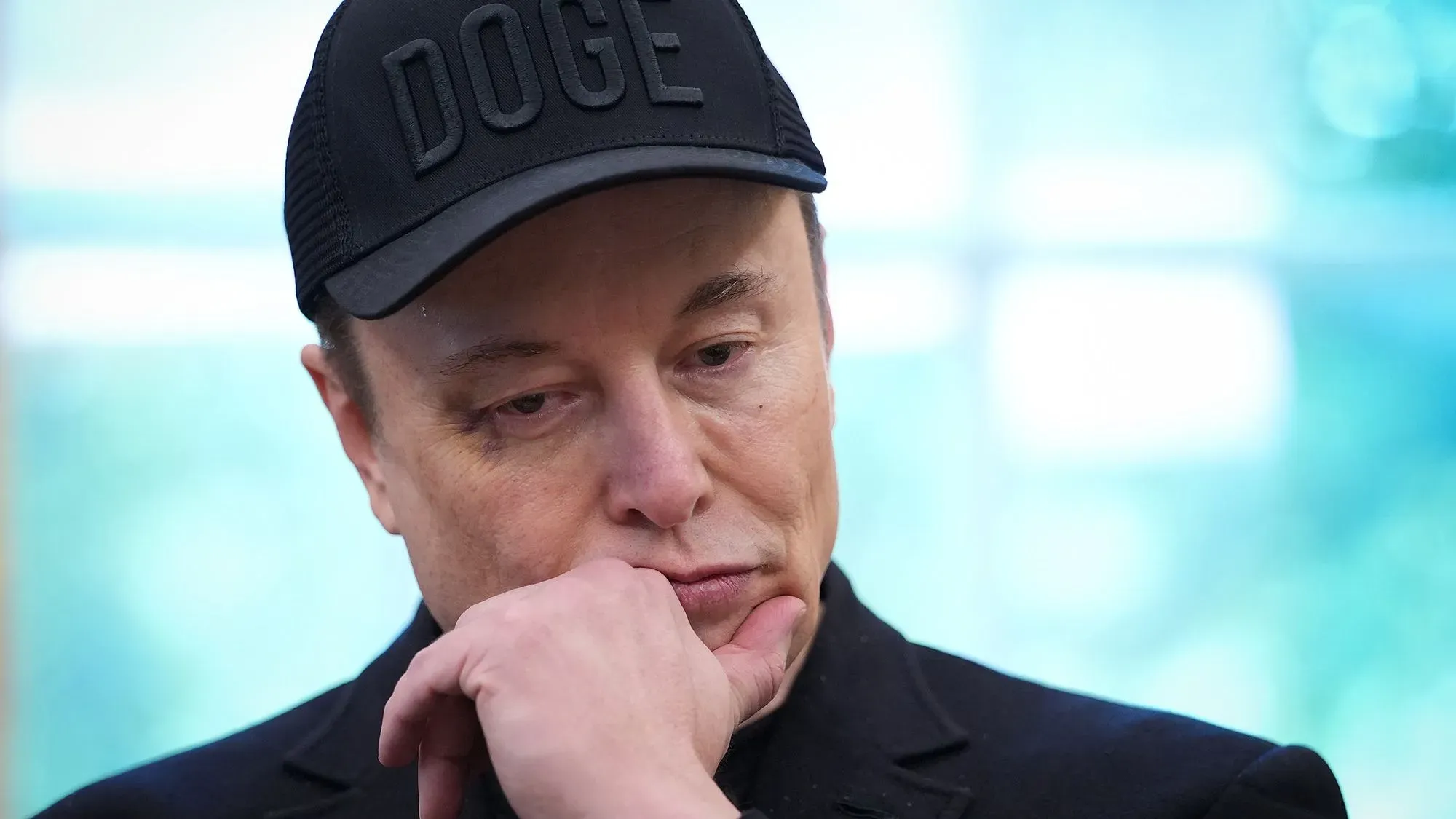 إيلون ماسك يبدو متأملاً وهو يرتدي قبعة تحمل شعار "DOGE"، مع خلفية ضبابية تعكس جوًا من القلق حول التشريعات السياسية.