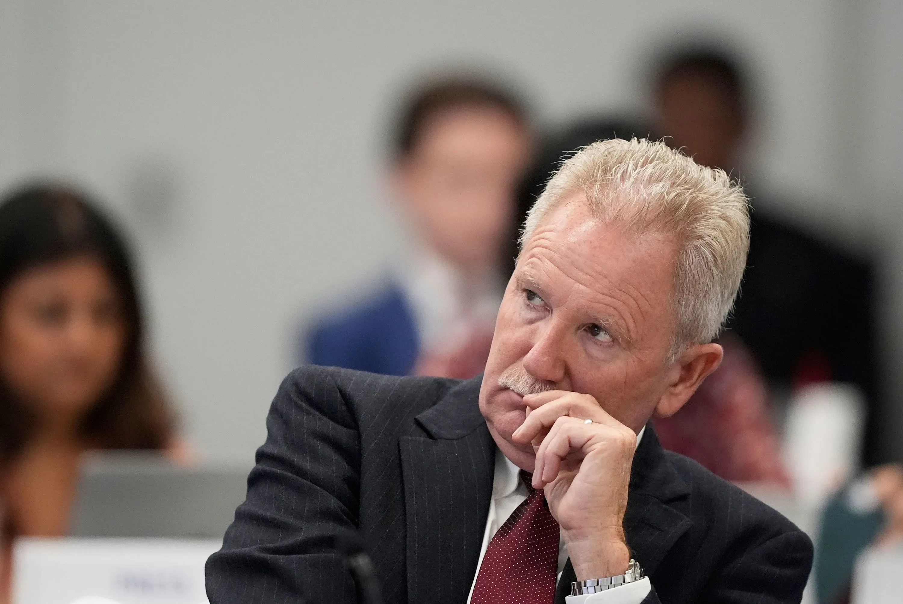 دكتور بروس كارلتون يستمع بتركيز خلال اجتماع حول سلامة لقاح كوفيد-19، مع وجود مشاركين آخرين في الخلفية.