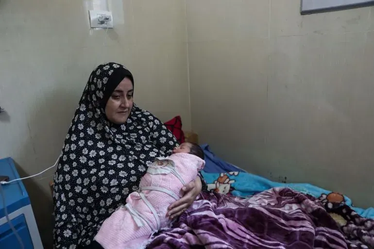 امرأة ترتدي حجابًا أسود تجلس على فراش في شقة مهدمة، تحمل مولودها الجديد بعد عملية قيصرية، تعكس معاناتها في ظل الظروف الصعبة في غزة.