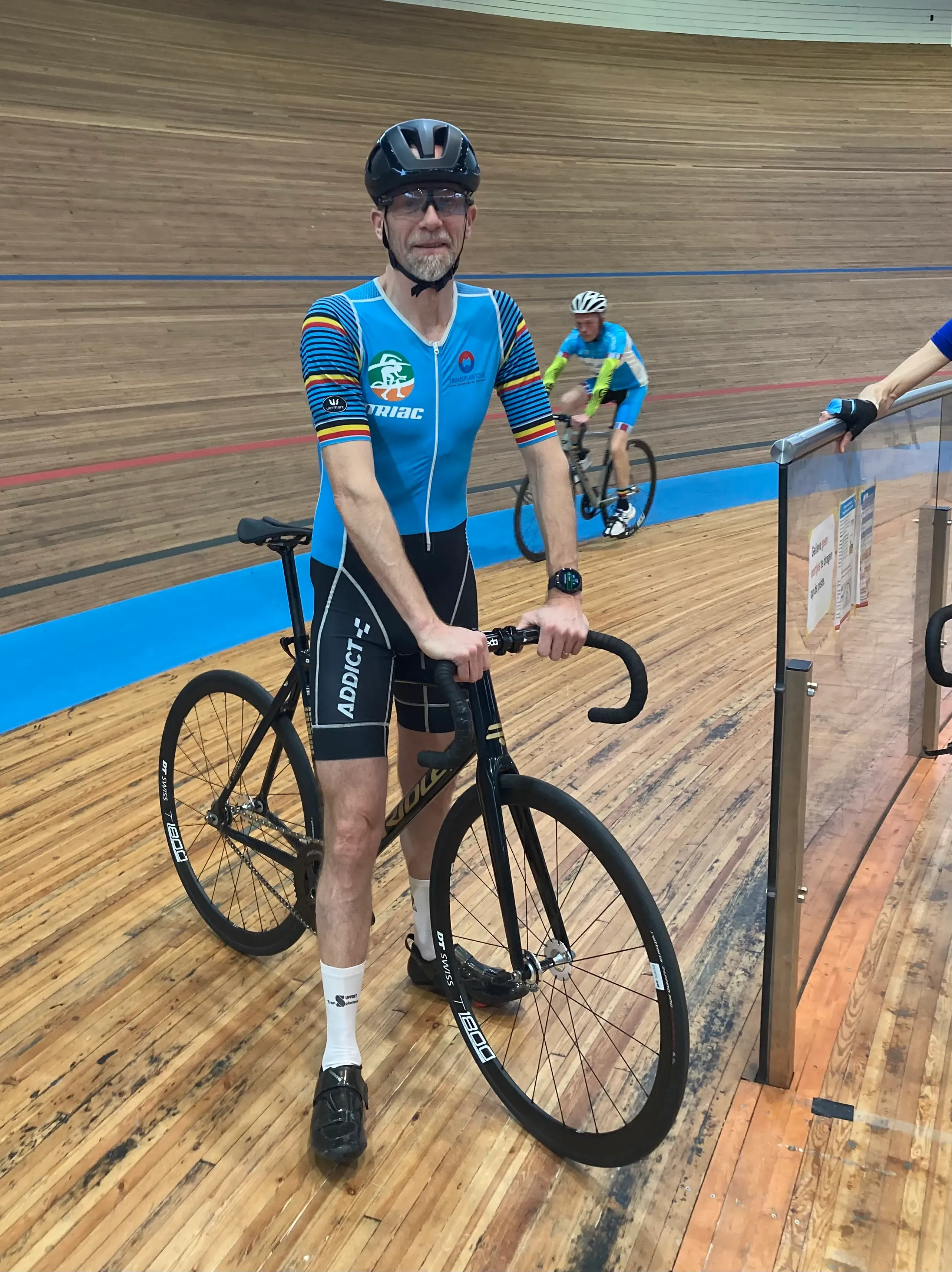 متسابق في دورة الألعاب العالمية لزراعة الأعضاء، يرتدي زي رياضي، يقف بجانب دراجته في مضمار خشبي، مع وجود متسابقين آخرين في الخلفية.