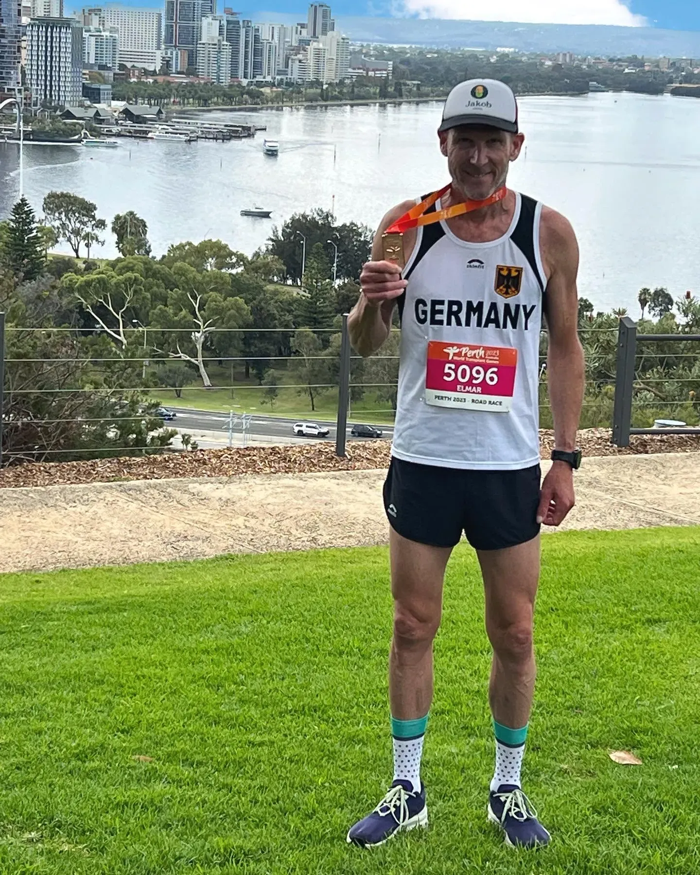 رياضي ألماني يحمل ميدالية ذهبية في سباق، يرتدي زي رياضي، ويقف أمام منظر خلاب لمدينة بيرث وأحد الأنهار.