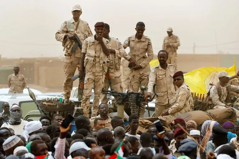 جنود من قوات الدعم السريع شبه العسكرية يقفون على مركبة عسكرية وسط حشد من المدنيين، مع وجود أسلحة ثقيلة في الخلفية، في سياق الصراع في شمال كردفان.