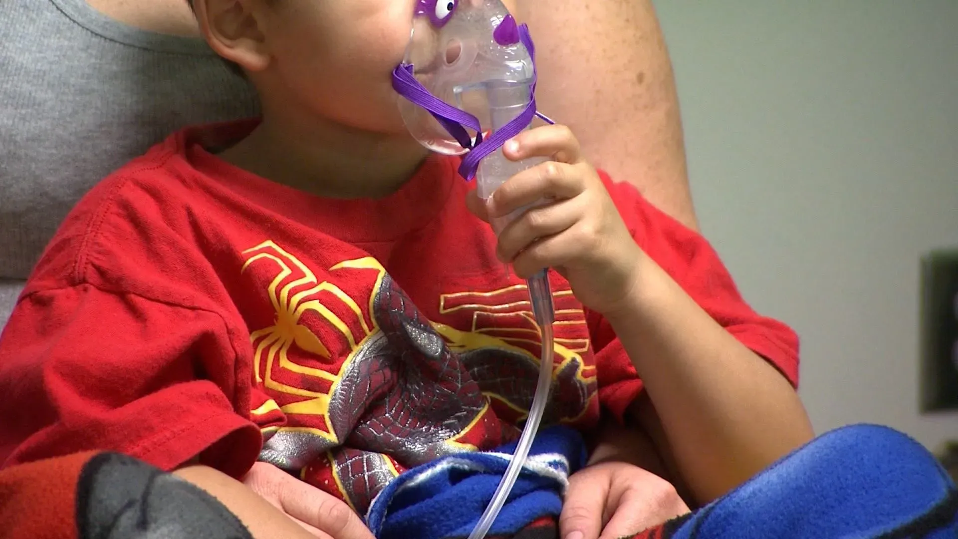 طفل يرتدي قميصًا أحمر ويستخدم جهاز استنشاق لعلاج فيروس RSV، يجلس في حضن شخص بالغ في بيئة طبية.