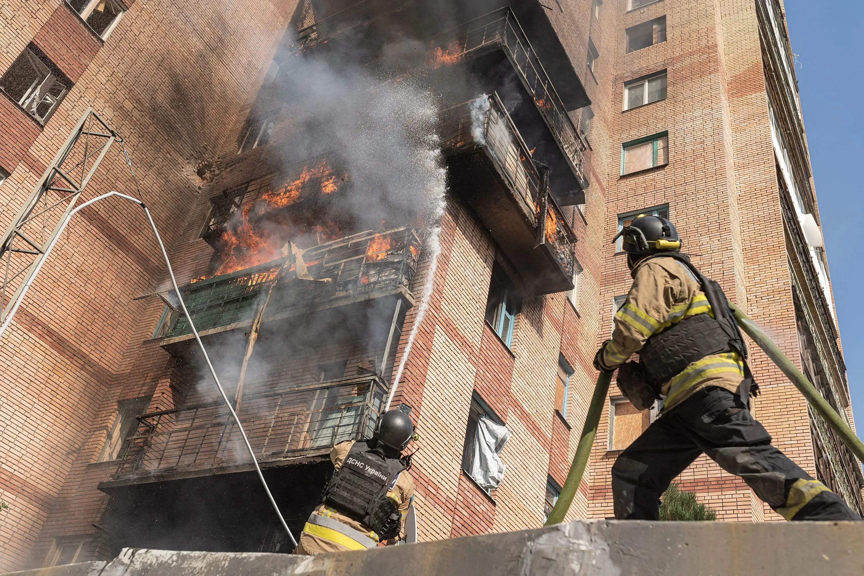 رجال الإطفاء يكافحون حريقًا في شقة بالطابق العلوي لمبنى سكني، حيث تتصاعد الأدخنة والنيران من الشرفة.