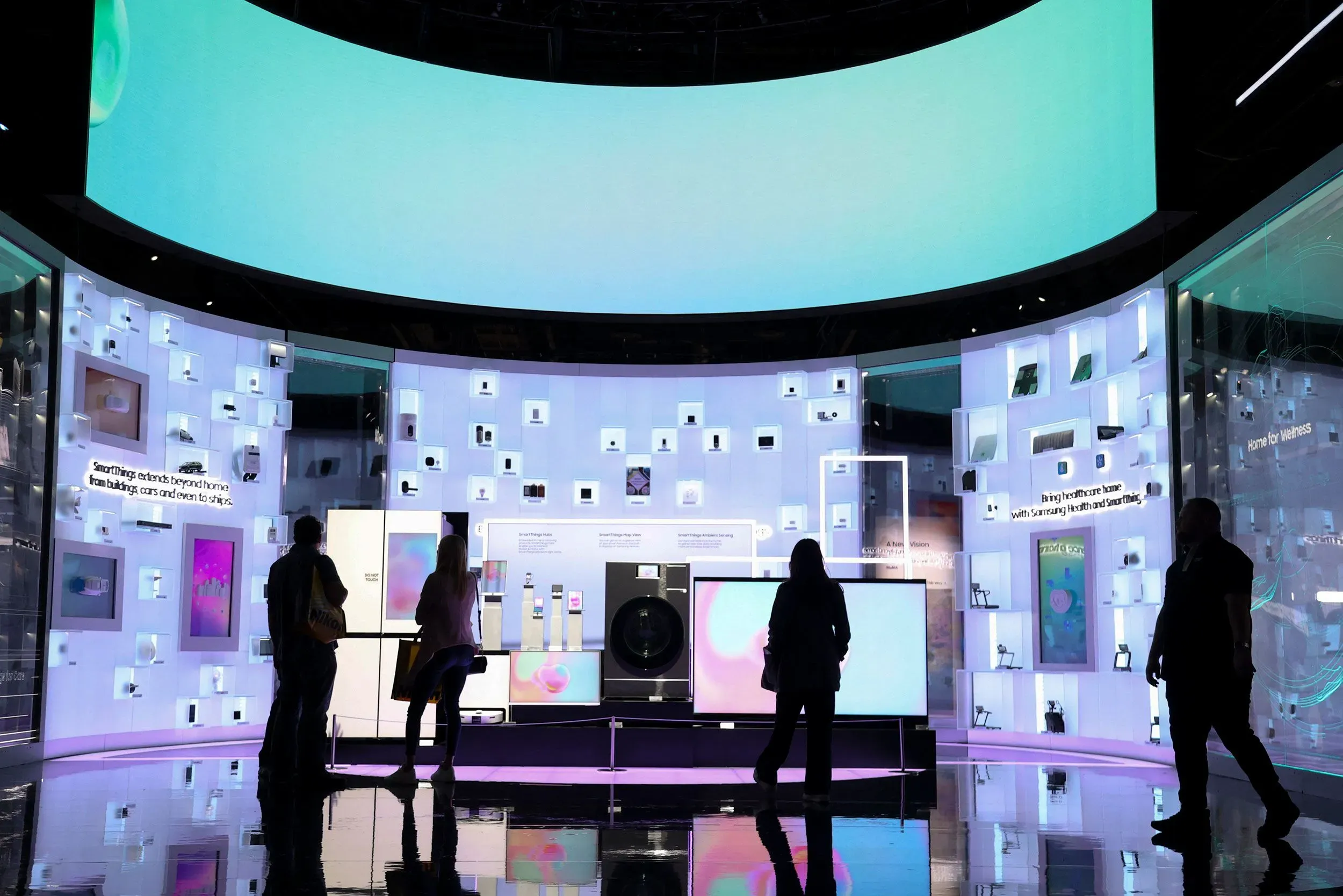 معرض تفاعلي لشركة سامسونج يظهر مجموعة من الأجهزة الذكية الحديثة، مع أشخاص يتفاعلون مع الشاشة الكبيرة والإضاءة الملونة.