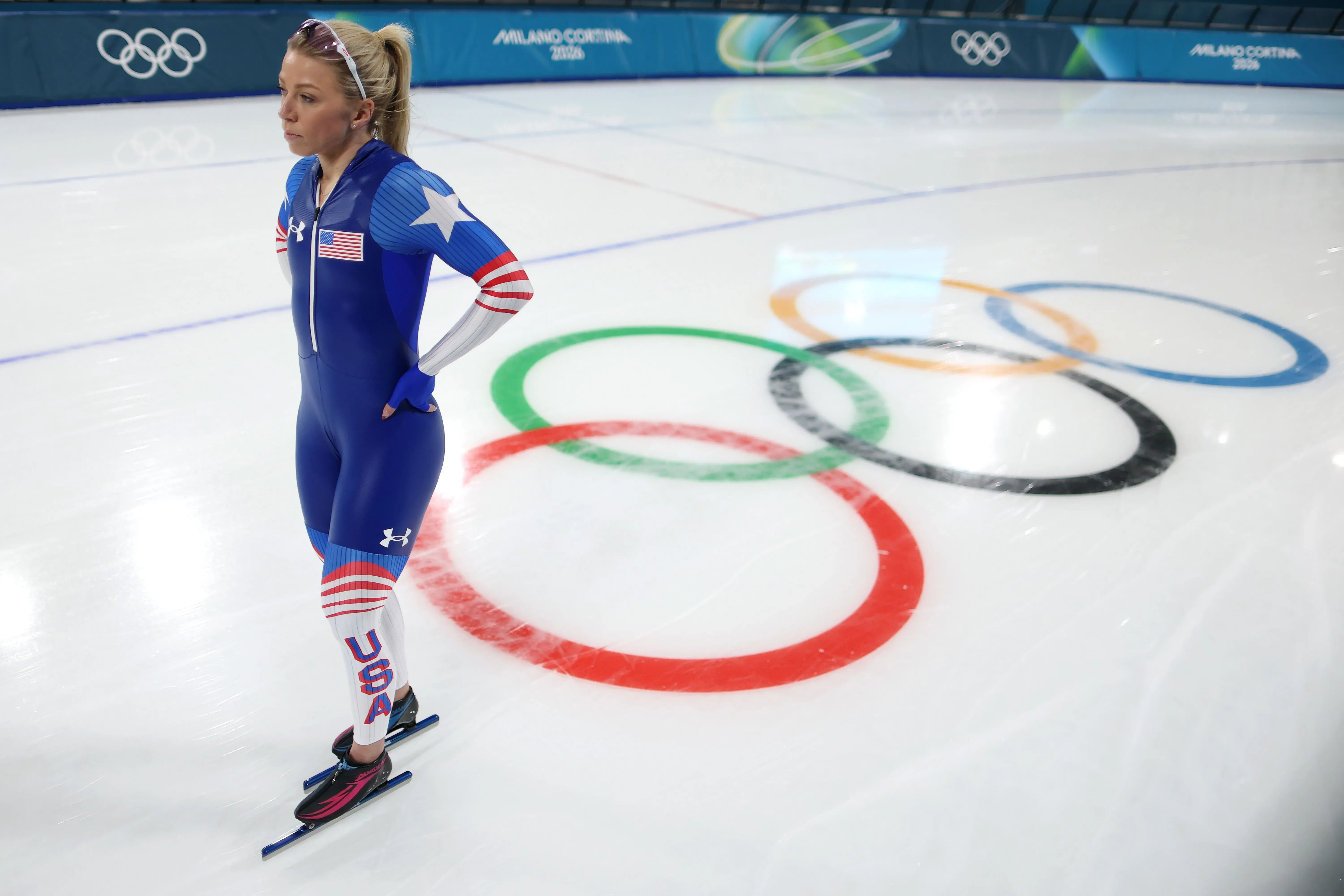سارة وارن ترتدي زي التزلج الأزرق مع شعار الولايات المتحدة، تقف على حلبة التزلج الأولمبية، مستعدة للمنافسة في سباق 500 متر.