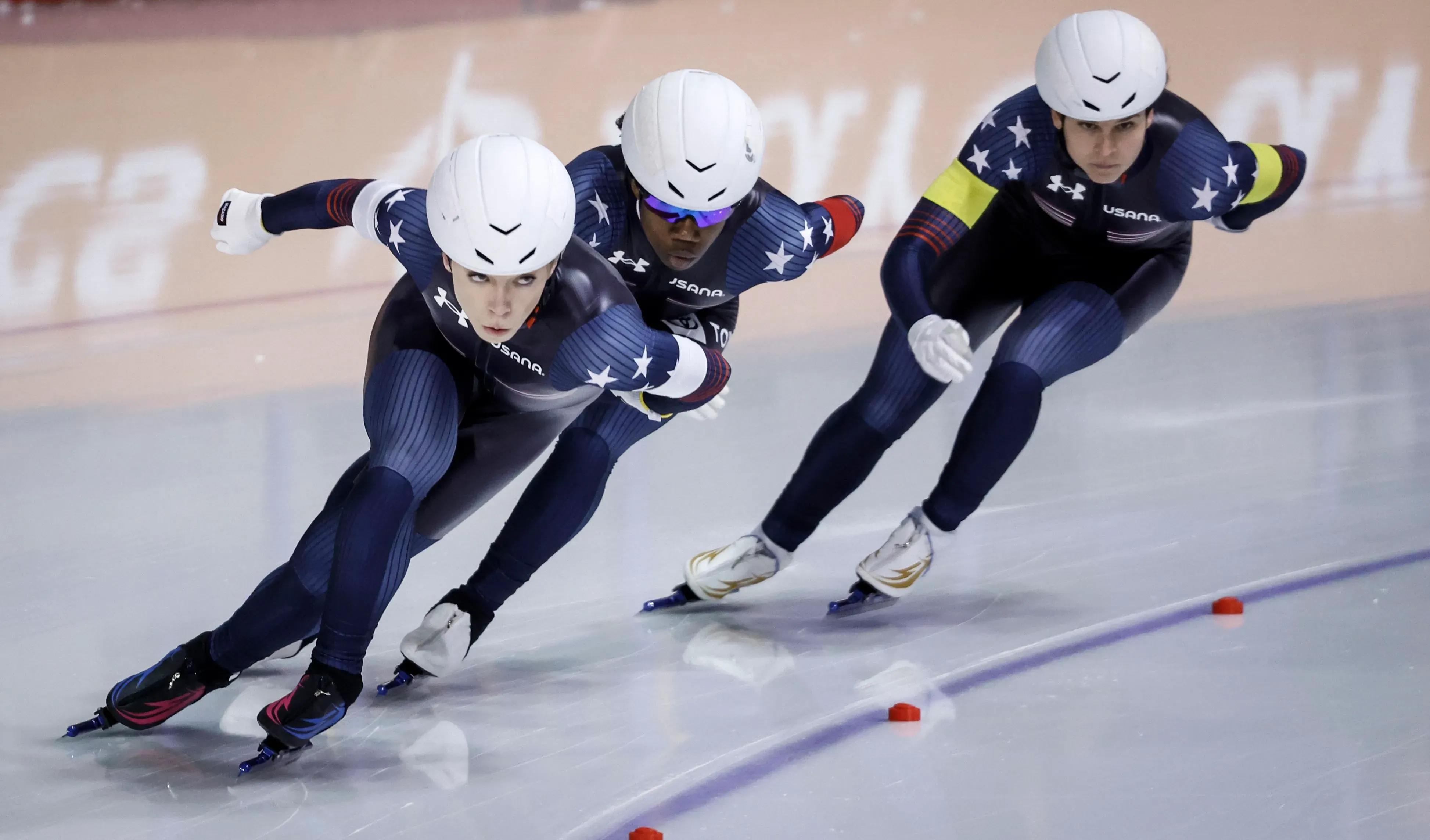 ثلاثة متزلجين يرتدون زي فريق الهوكي الأمريكي يتسابقون في حلبة التزلج، مع التركيز على الحركة الديناميكية والتنافس في سباق التزلج السريع.