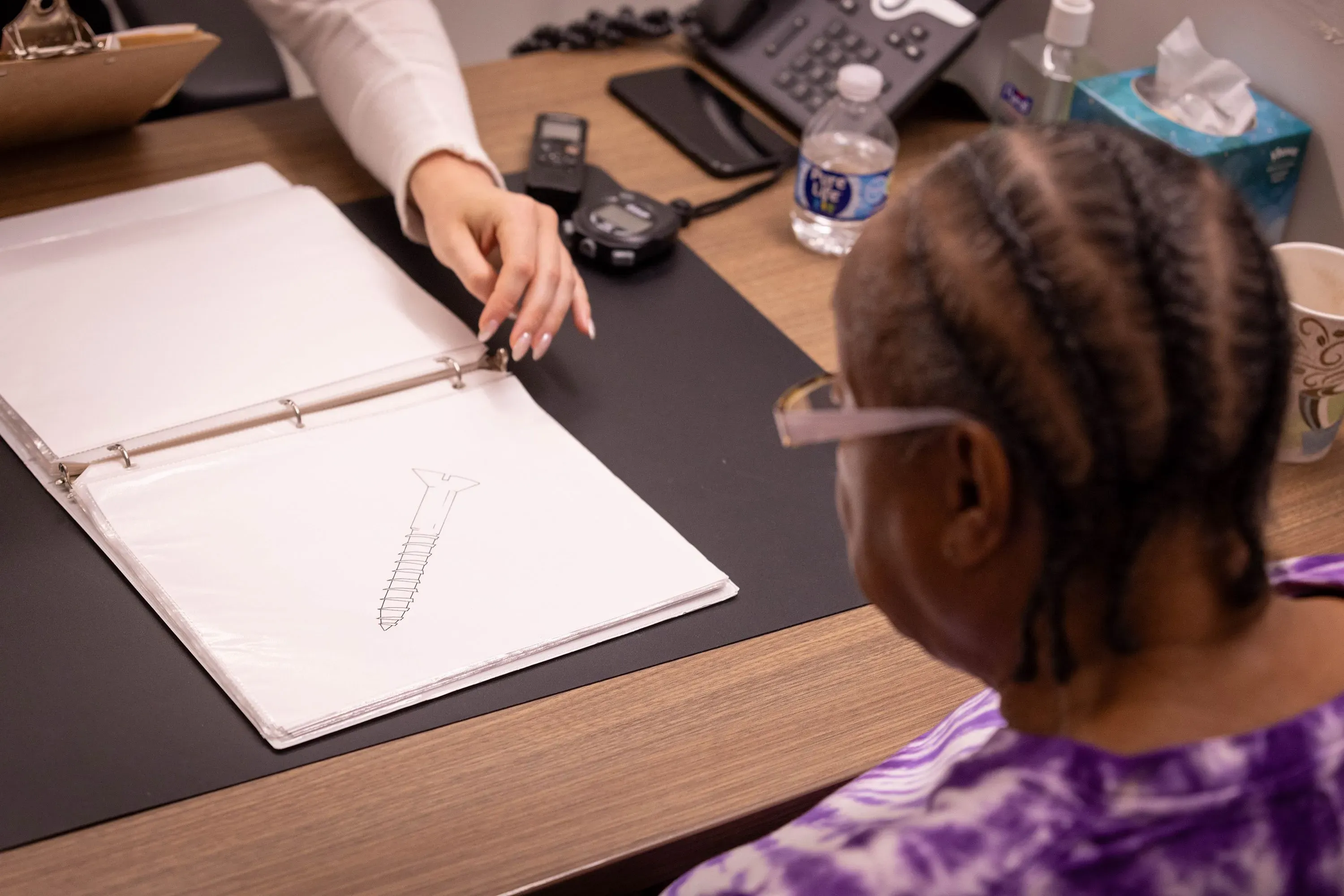 امرأة مسنّة ترتدي نظارات، تنظر إلى رسم توضيحي لمسمار، بينما تشير إليه طبيبة خلال جلسة تقييم إدراكي.