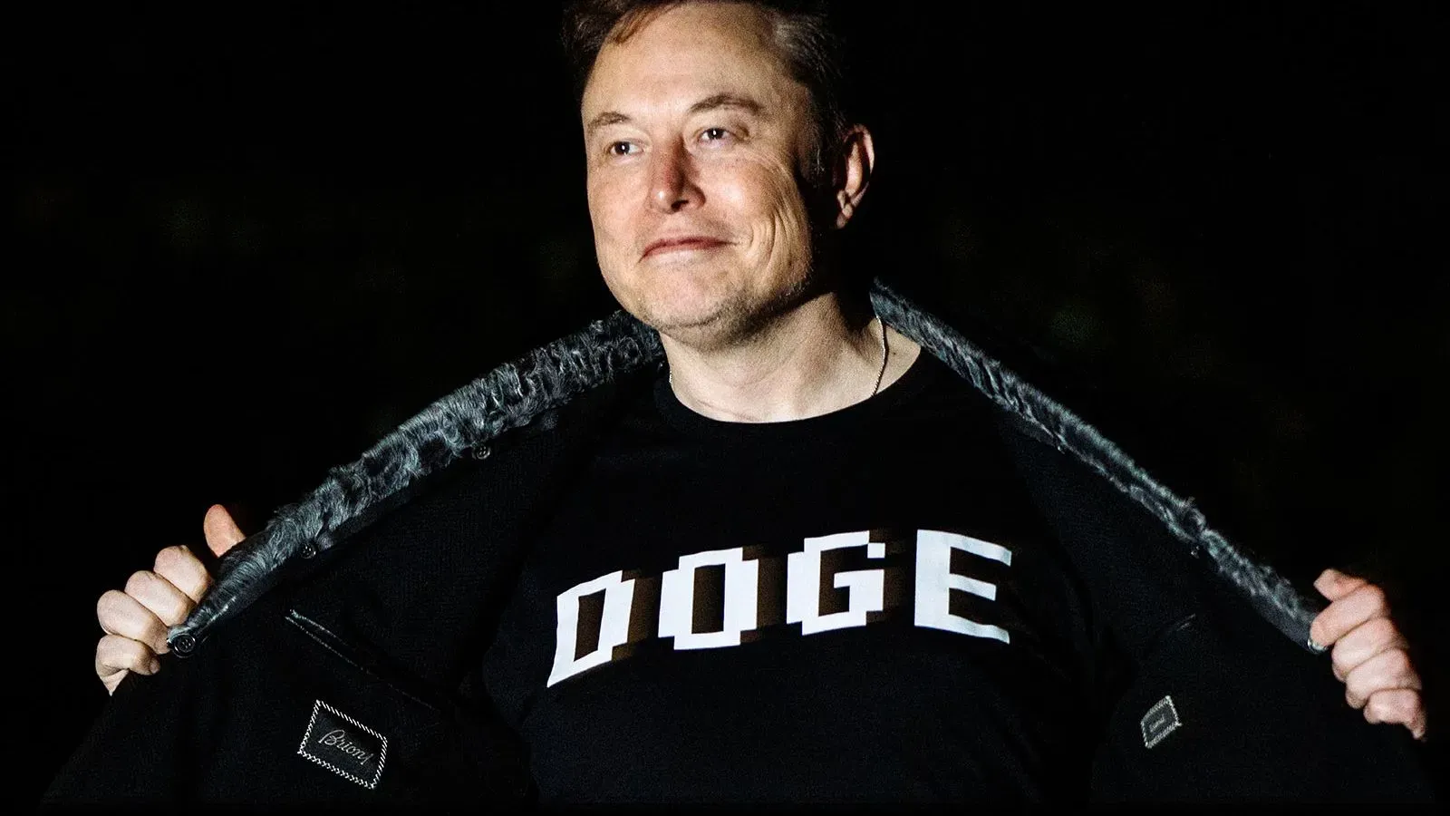 إيلون ماسك يرتدي قميصًا مكتوبًا عليه "DOGE"، مبتسمًا في صورة تظهره في بيئة مظلمة، تعكس تأثيره على السياسة والاقتصاد.