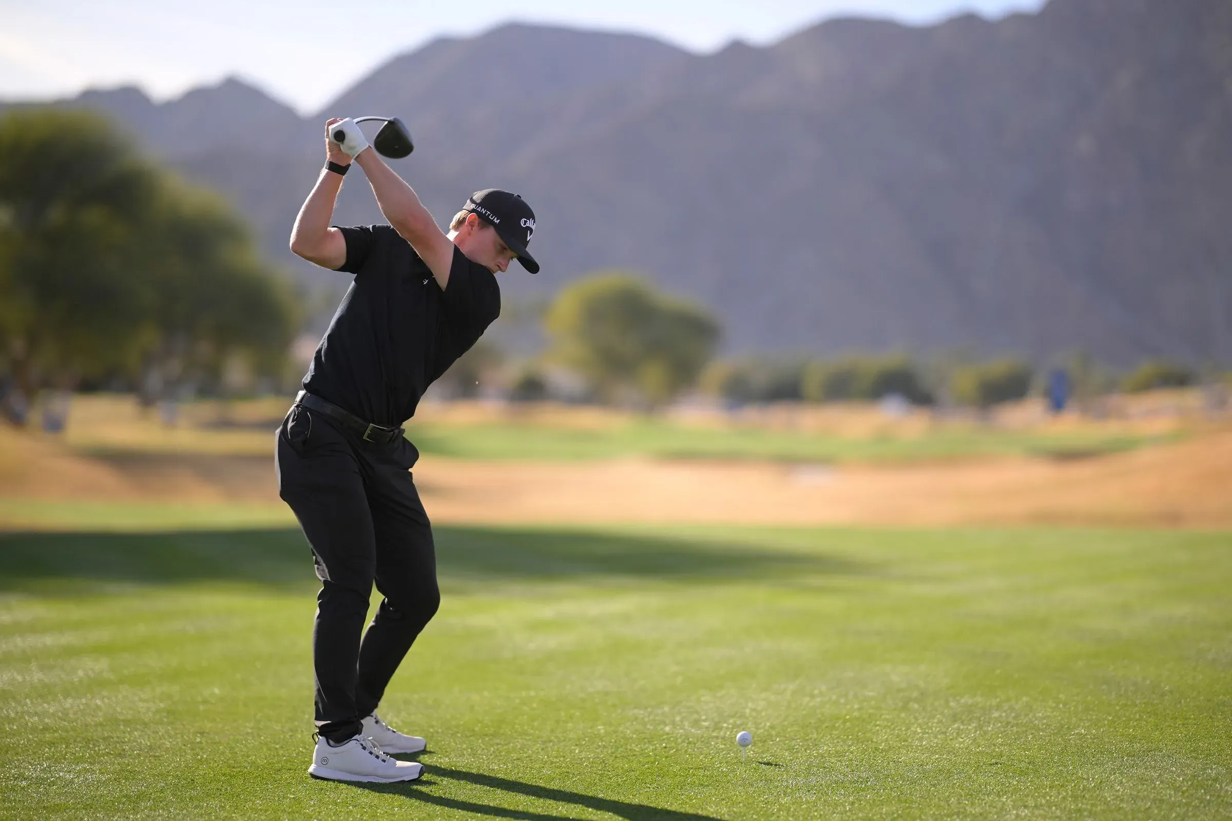 لاعب غولف شاب يرتدي ملابس سوداء، يستعد لضرب الكرة في ملعب غولف محاط بالجبال، مع التركيز على أدائه في بطولة أمريكان إكسبريس.