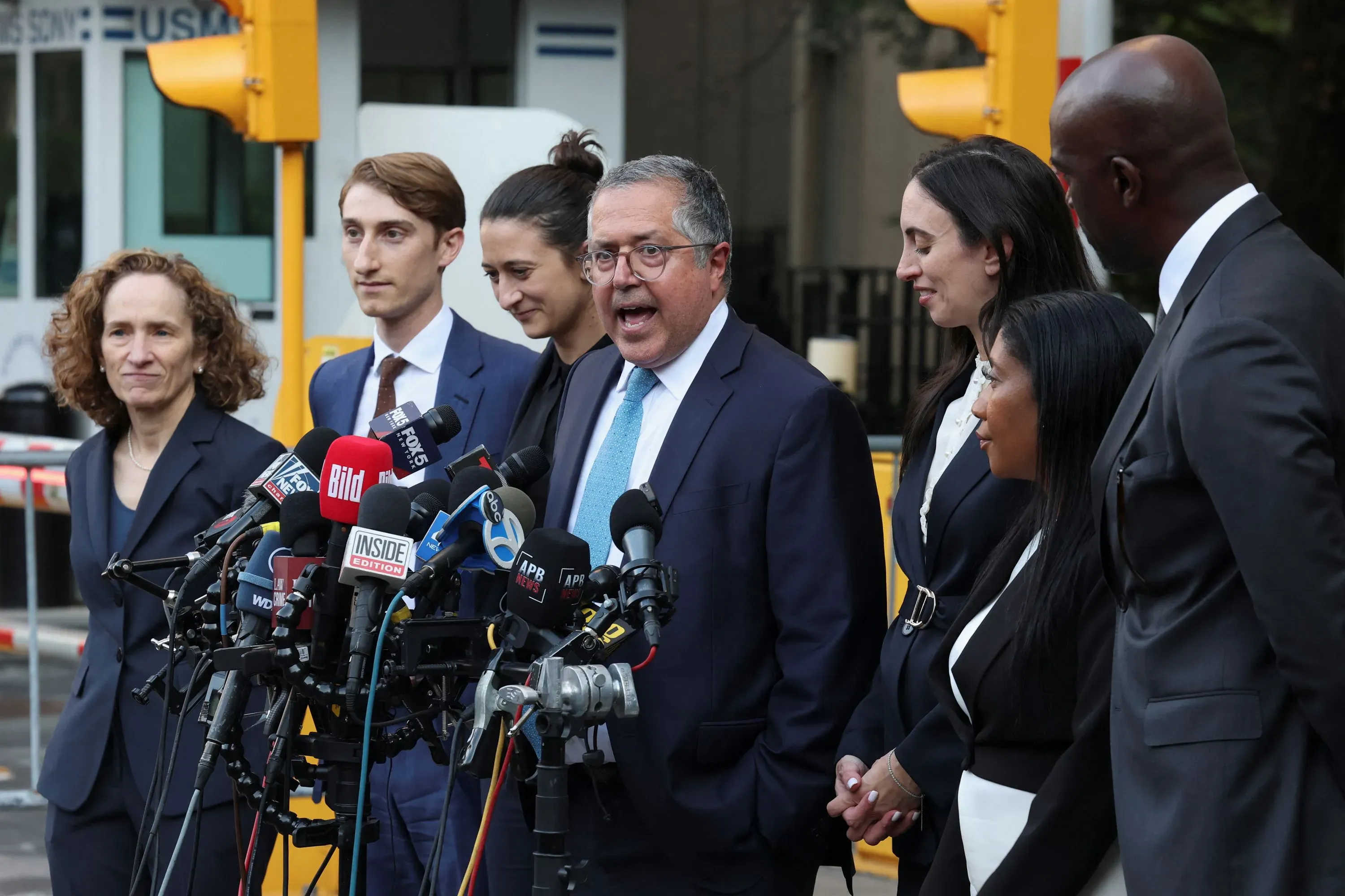 مجموعة من المحامين يتحدثون أمام وسائل الإعلام بعد جلسة استماع تتعلق بشون كومبس، مع وجود ميكروفونات وصحفيين في الخلفية.