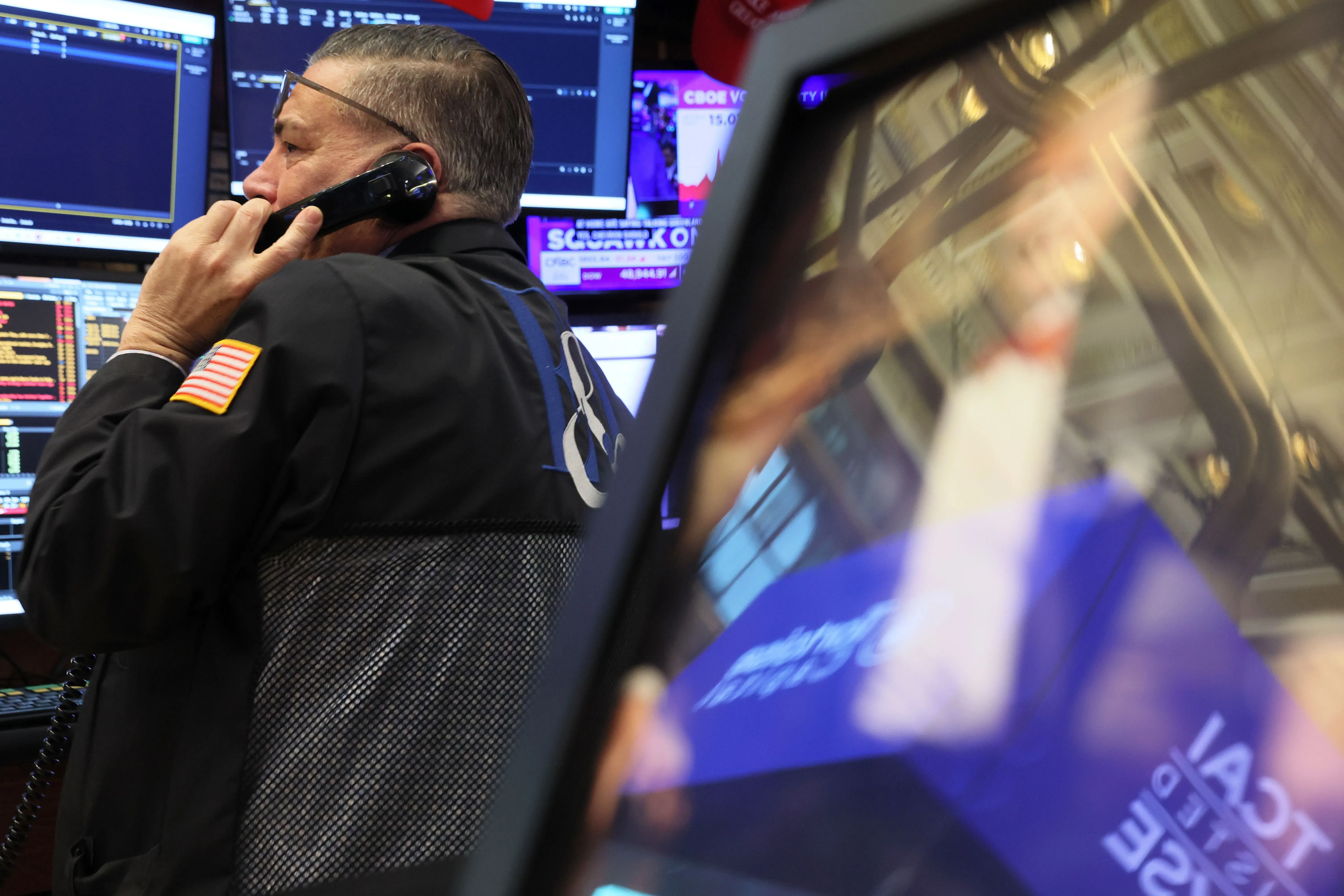 تاجر في بورصة الأسهم يتحدث عبر الهاتف، محاط بشاشات تعرض بيانات السوق، مع تراجع ملحوظ في الأسهم والدولار.