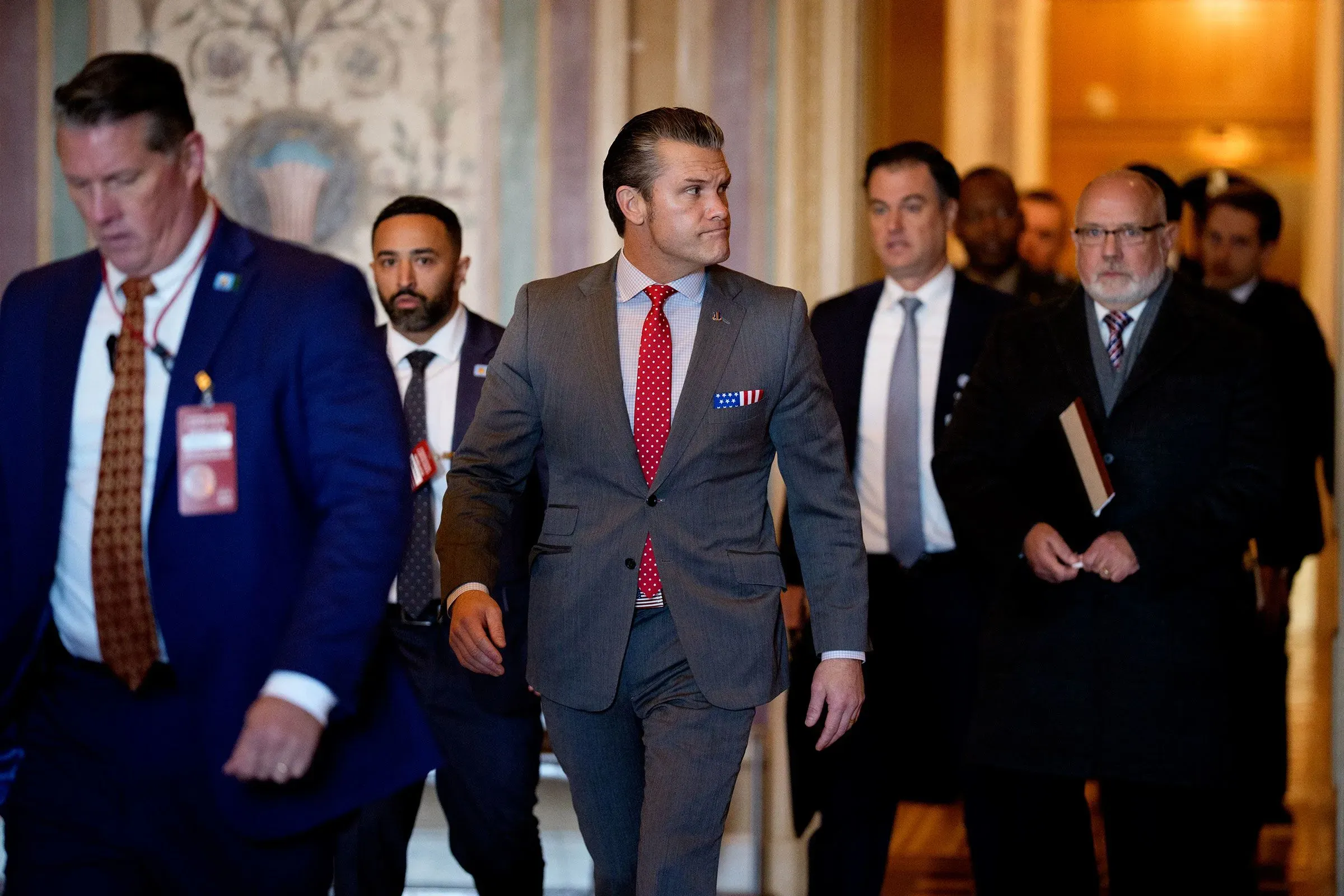 مجموعة من السياسيين يسيرون في مبنى مجلس الشيوخ، بينهم رجل يرتدي بدلة رسمية مع شارة عسكرية، في سياق مناقشة قانون السياسة الدفاعية.