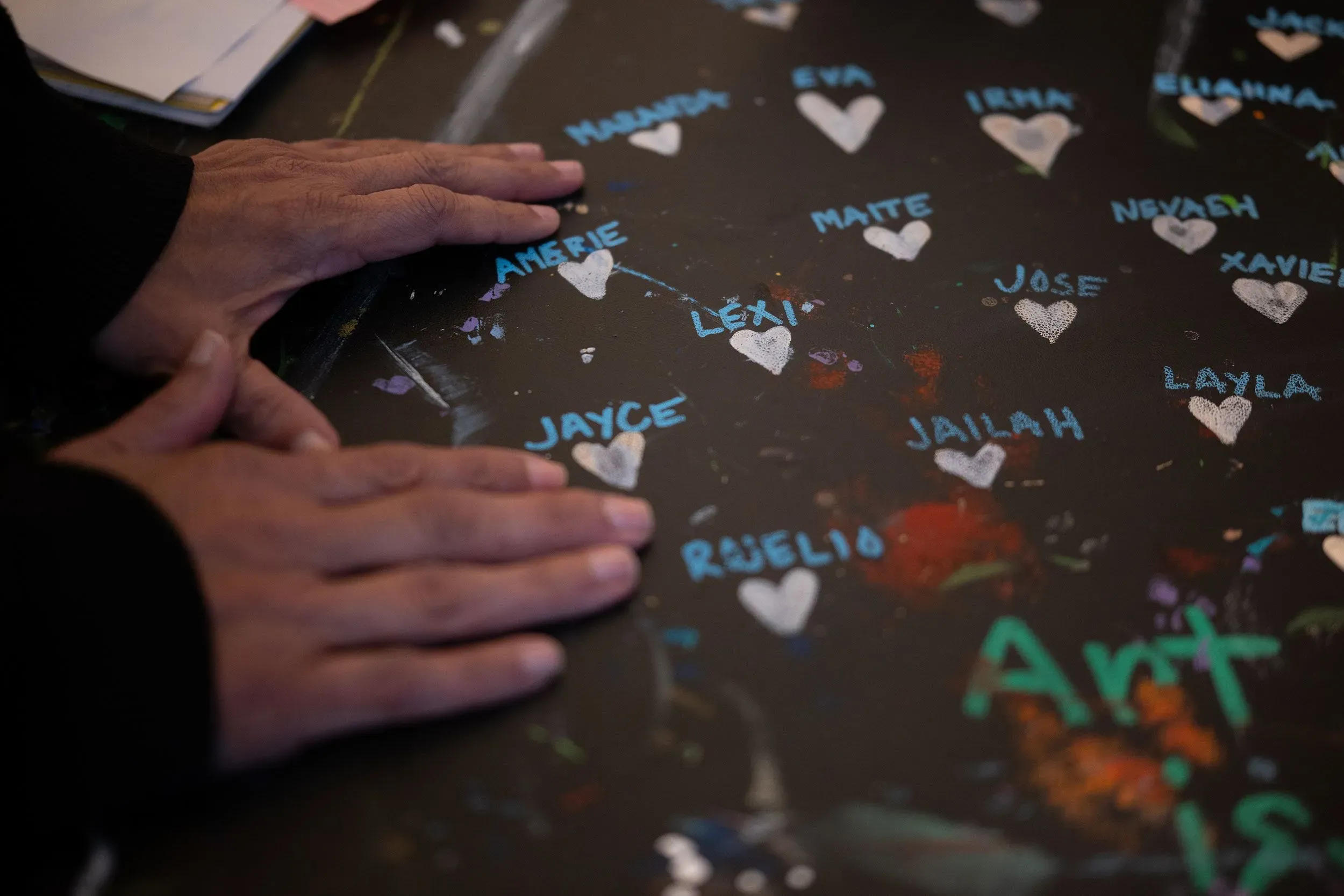 أيدٍ تلامس سطحًا مزينًا بأسماء أطفال قُتلوا، مع قلوب مرسومة حول الأسماء، تعبيرًا عن الحزن والذكرى.