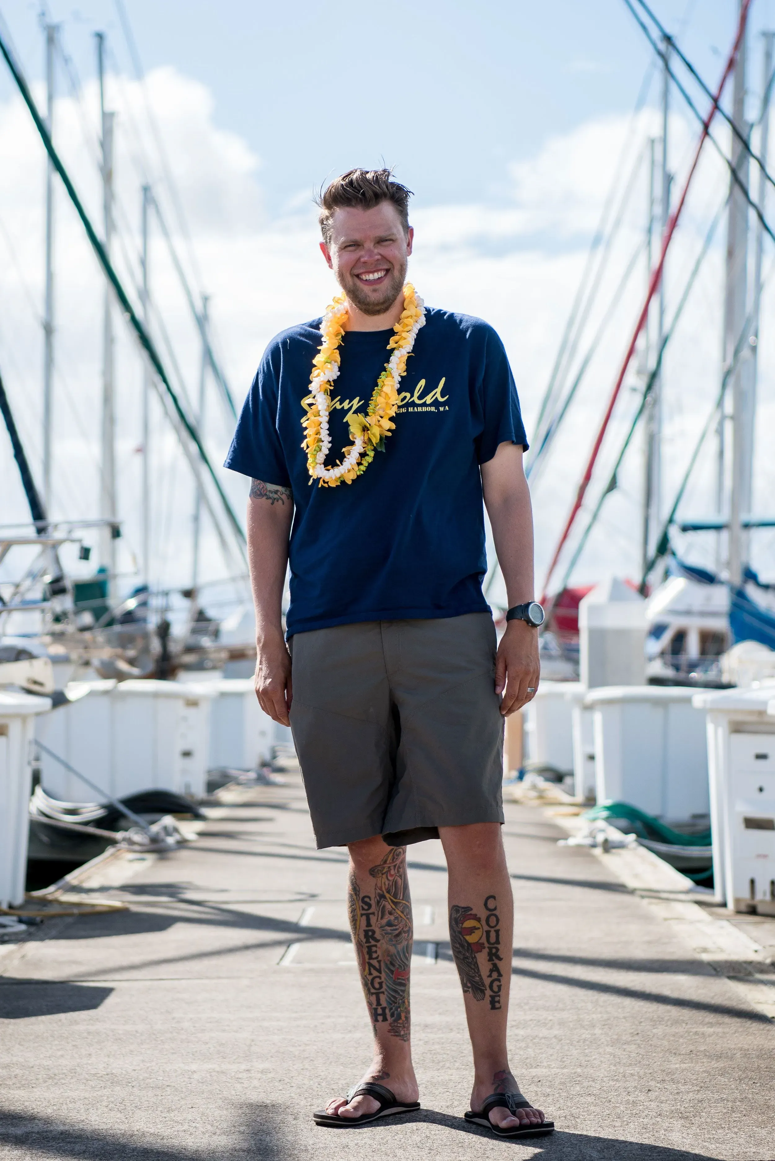 رجل مبتسم يرتدي قلادة زهور وشورتات قصيرة، يقف على رصيف بالقرب من قوارب في هونولولو، هاواي، مع خلفية بحرية.
