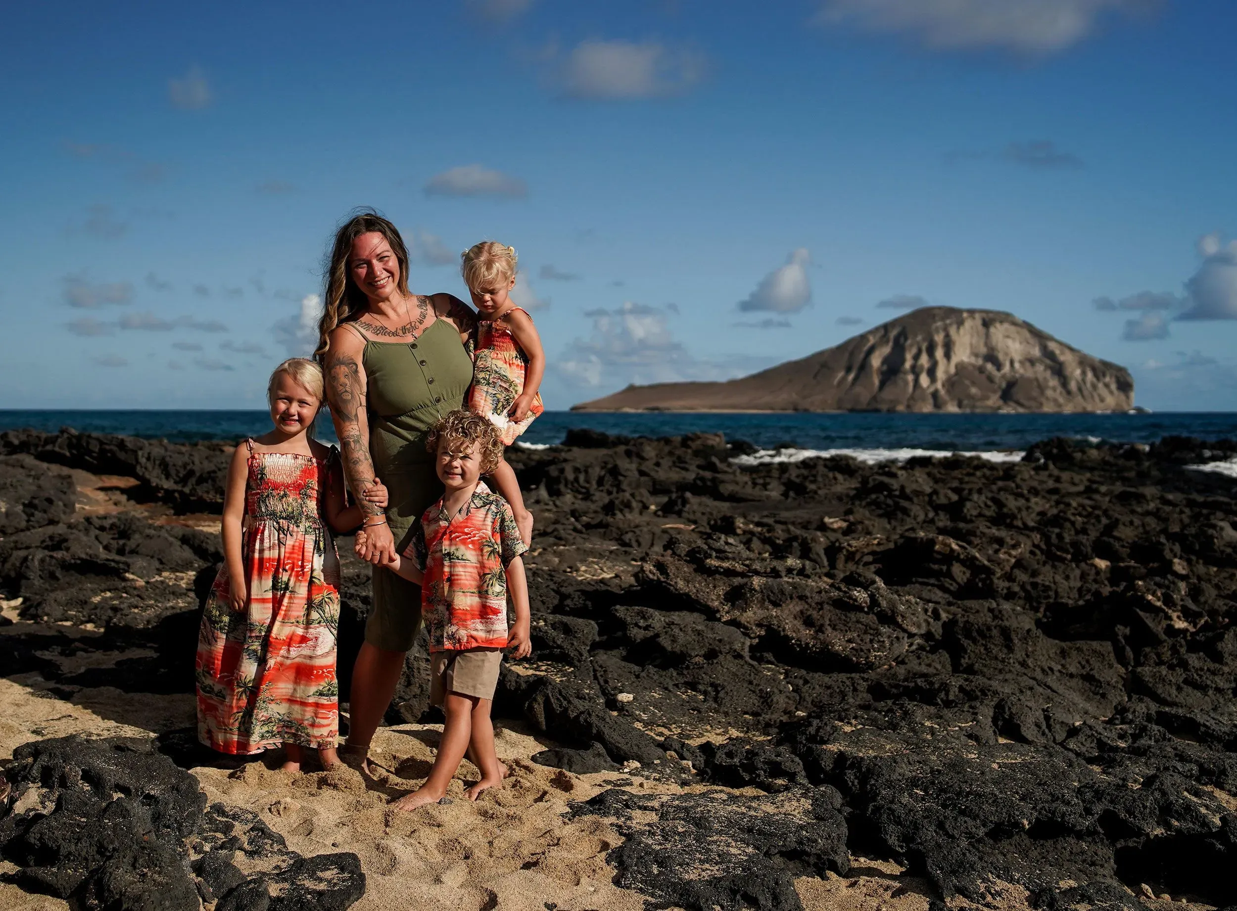 عائلة تتكون من أم وطفلين على شاطئ صخري في هاواي، مع منظر لجزيرة صغيرة في الخلفية، تعكس لحظات السعادة والذكريات.