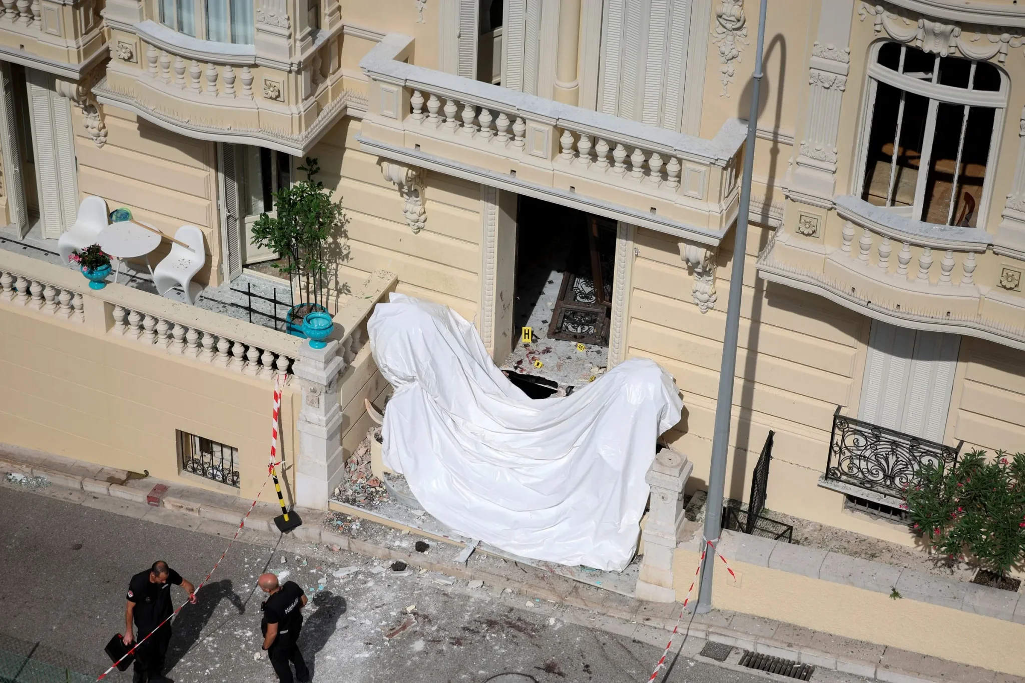 مدخل مبنى فاخر في موناكو تعرض لانفجار قنبلة، مع وجود شرطة تحقق في موقع الحادث وتأثيرات الدمار حول المدخل.