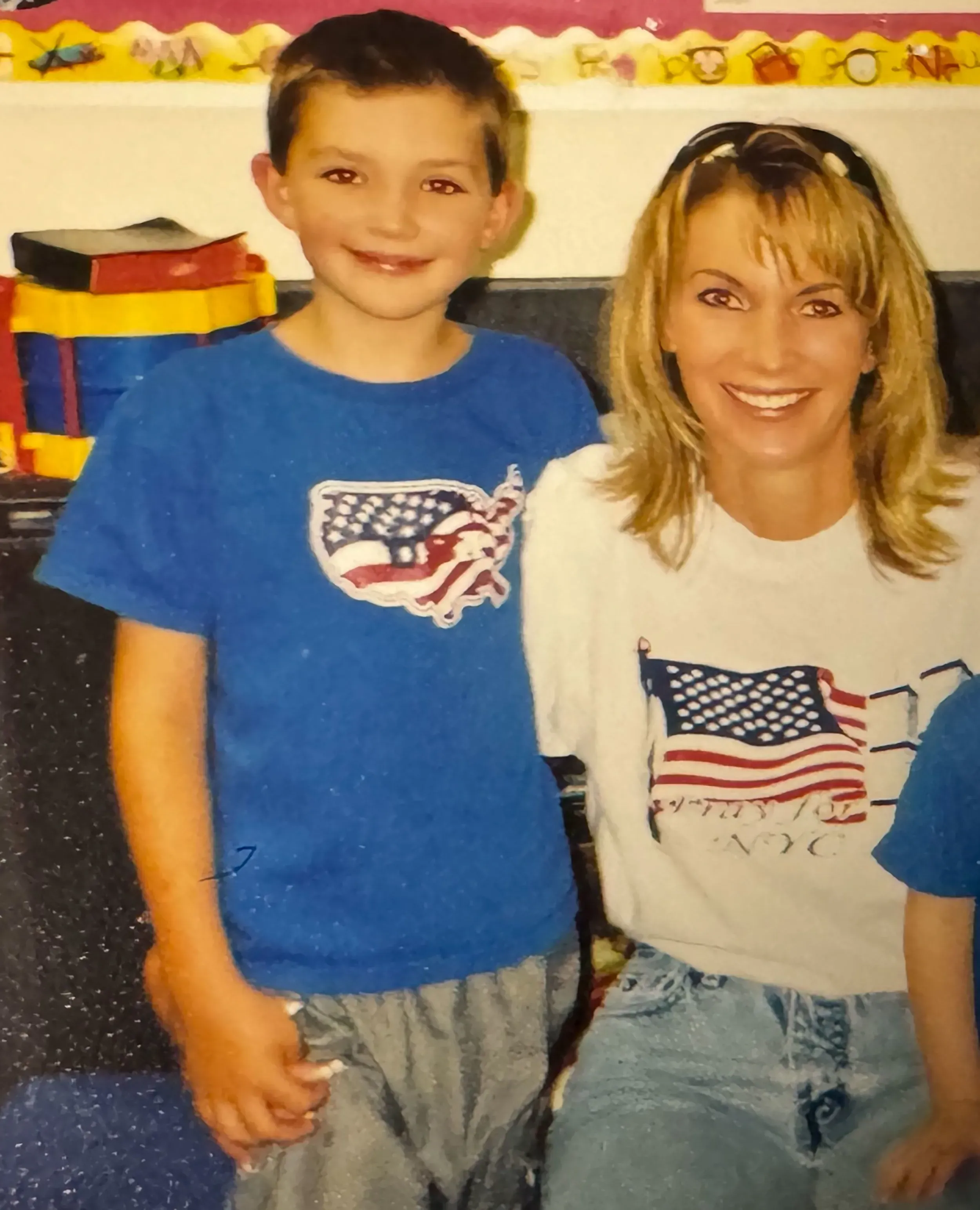 صورة تظهر أمًا مبتسمة مع طفل صغير يرتدي قميصًا أزرق، في خلفية صف دراسي ملون، تعكس لحظة من السعادة قبل أحداث مأساوية.