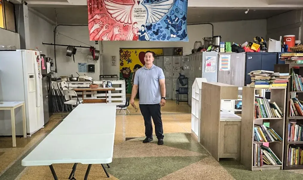 رجل يقف في مركز مجتمعي في تيخوانا، محاط بمكتبة وأثاث بسيط، يعكس جهود دعم المهاجرين وتحقيق الأحلام الجديدة.