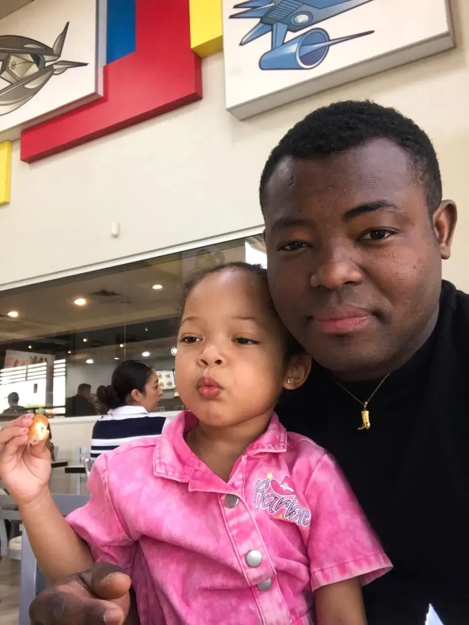 رجل مع ابنته الصغيرة في مطعم، حيث تبدو الفتاة ترتدي قميصًا ورديًا وتتناول الطعام، بينما يظهر خلفهم بعض الزبائن.