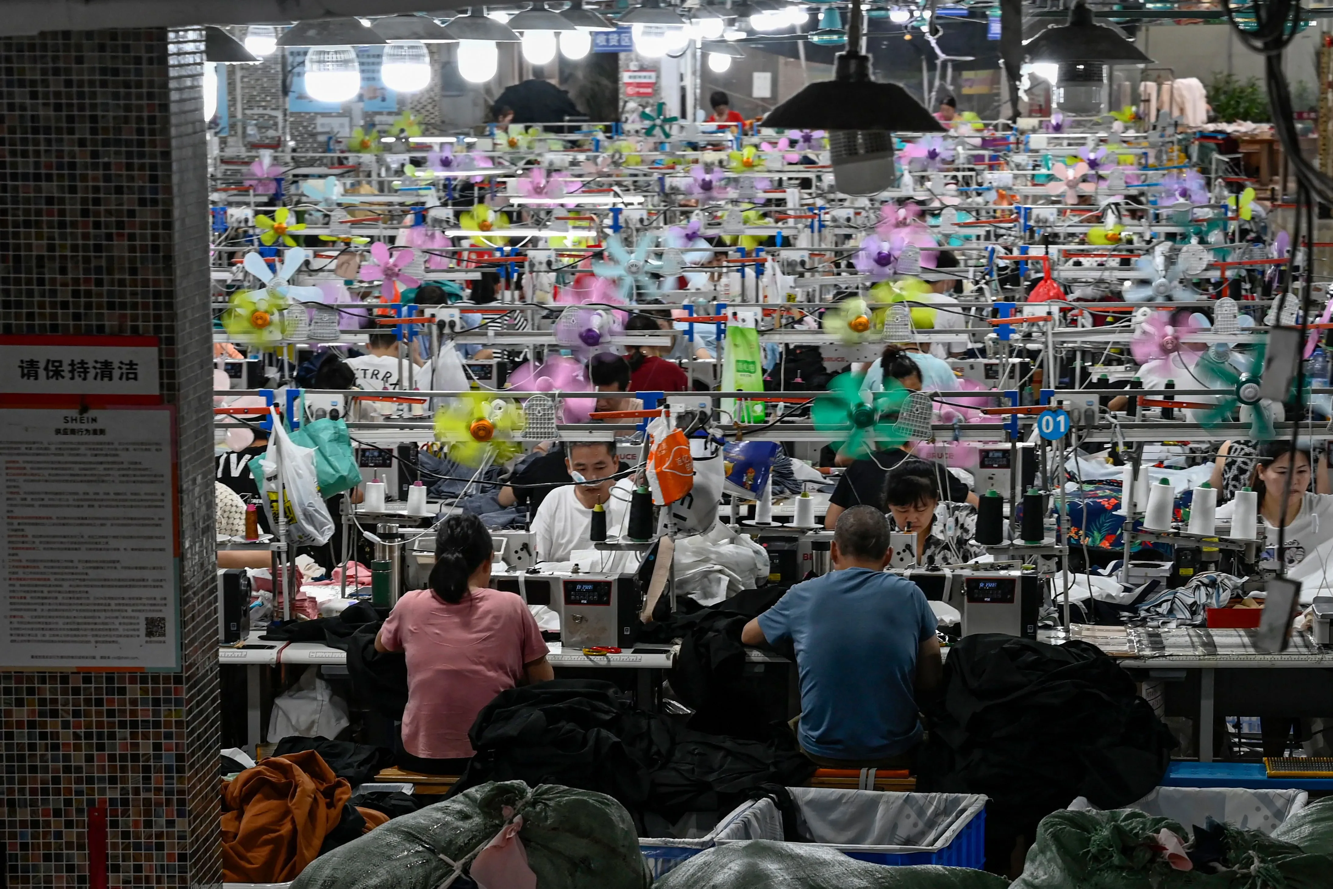 صورة لعمال في مصنع ملابس، حيث يظهر العديد منهم يعملون على آلات الخياطة وسط زينة ملونة، مما يعكس تأثير التجارة العالمية.