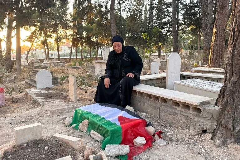 امرأة فلسطينية ترتدي الحداد تجلس بجانب قبر مغطى بعلم فلسطين في مقبرة بمخيم دير عمّار بالضفة الغربية المحتلة.