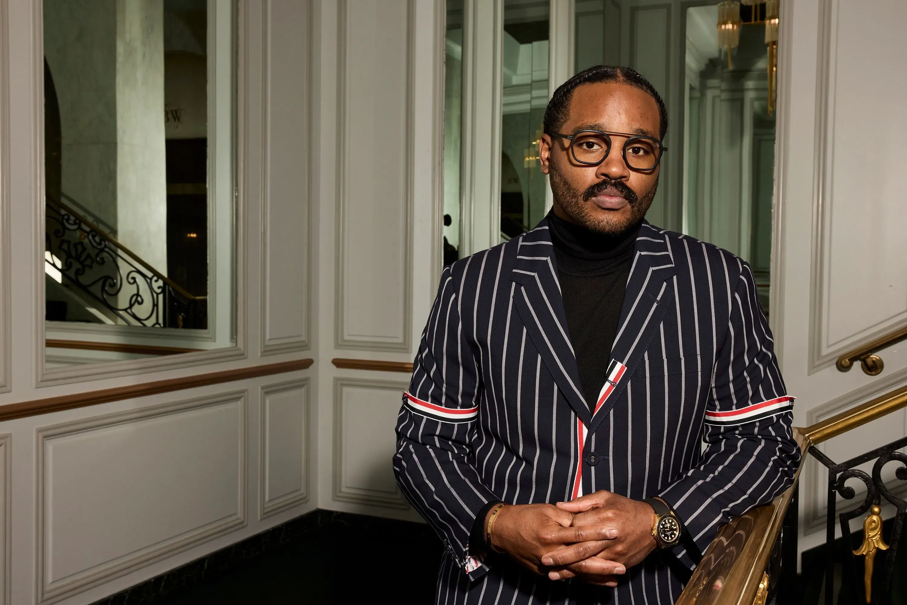 صورة لرجل يرتدي بدلة مخططة، يقف بجانب درابزين في مكان أنيق، يعكس نجاحه في صناعة السينما وتأثيره الثقافي.