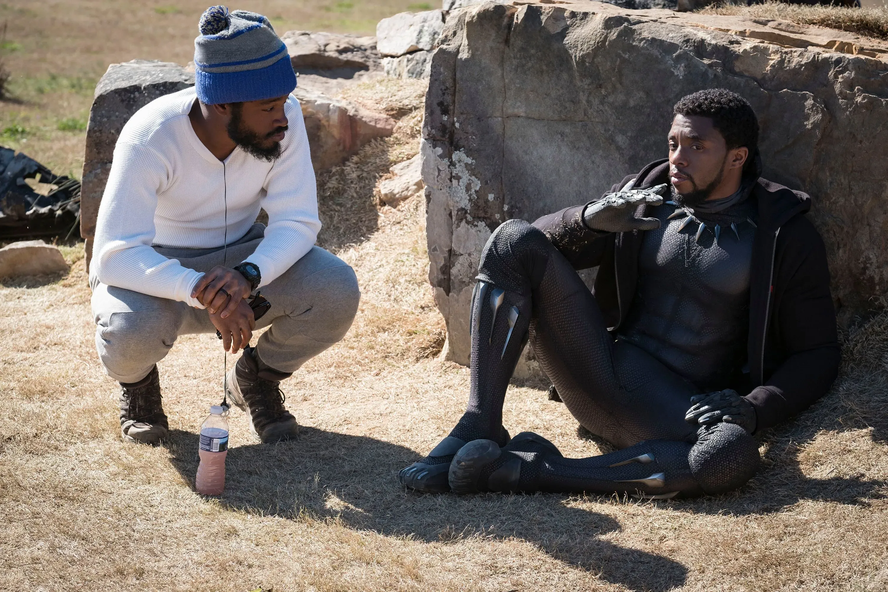 مشهد من فيلم "Black Panther" يظهر شخصيتين، أحدهما جالس على الأرض يرتدي زي البطل، بينما الآخر يستمع بانتباه.
