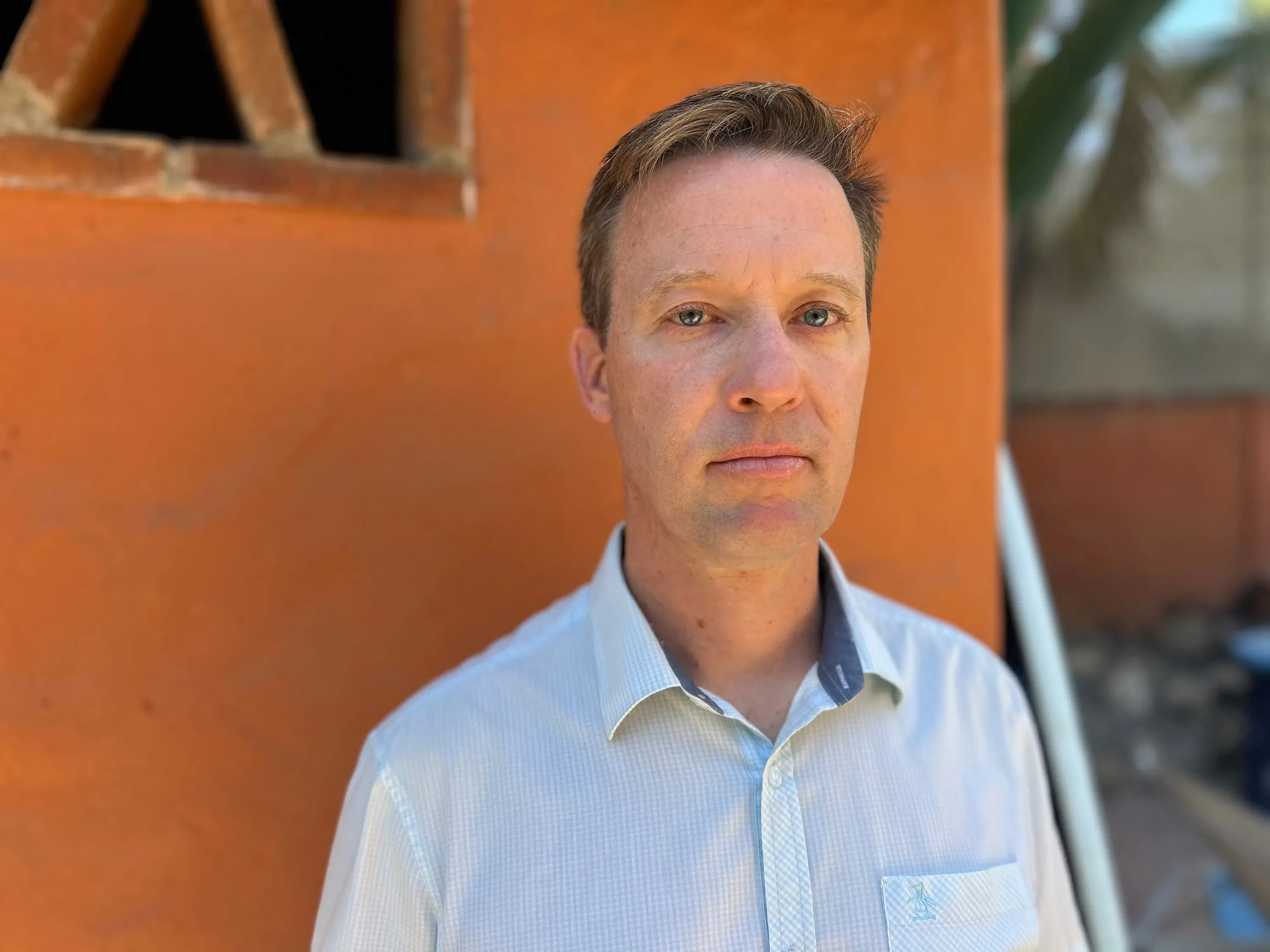 عالم في مجال الأبحاث الطبية يقف أمام جدار برتقالي، معبرًا عن القلق بشأن تأثيرات إدارة ترامب على تمويل الأبحاث العلمية.