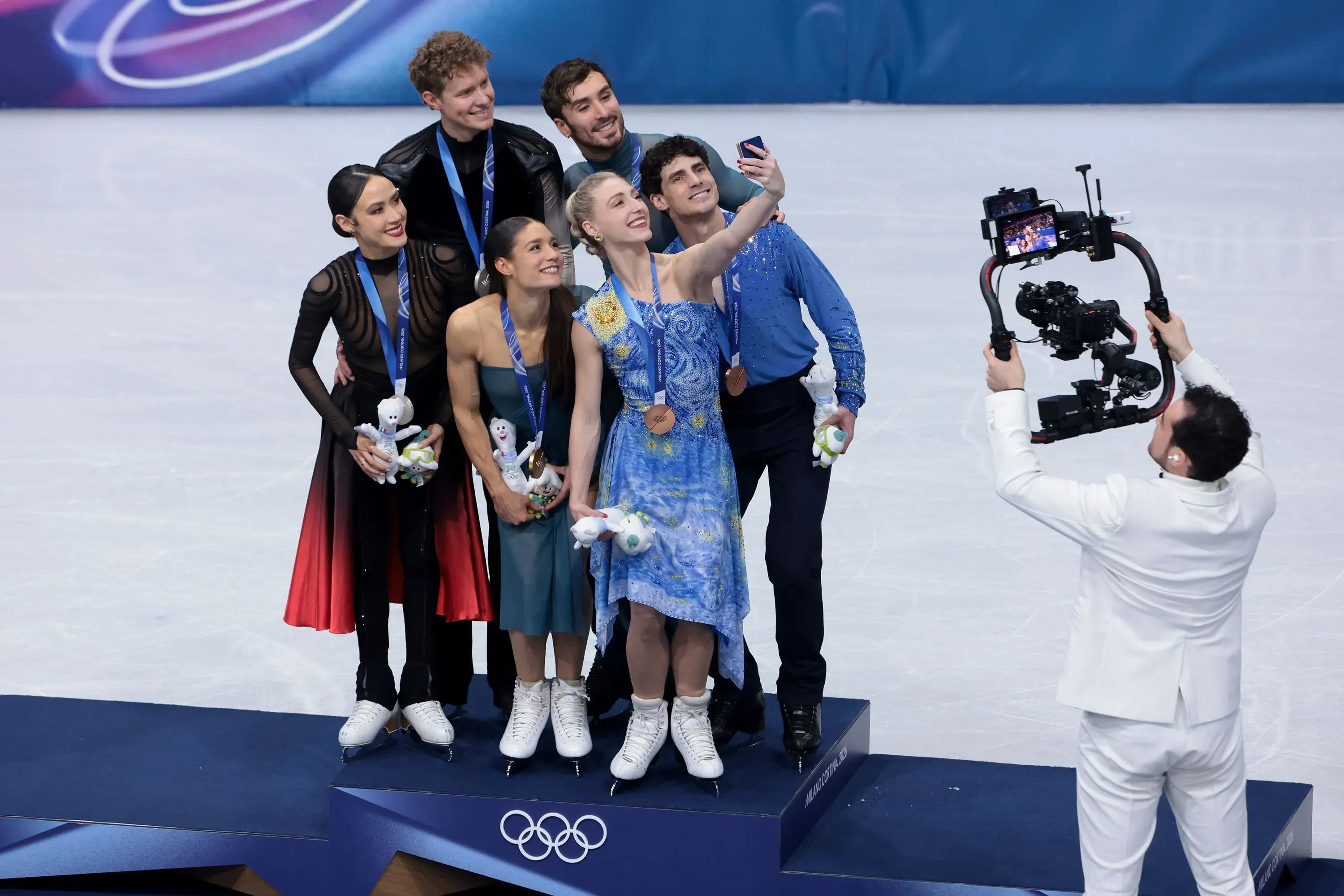 جوردان كوان، المصور الأولمبي، يلتقط صورة سيلفي مع المتزلجين الفائزين بالميداليات على الجليد في ميلانو، مرتديًا بدلة تزلج بيضاء.